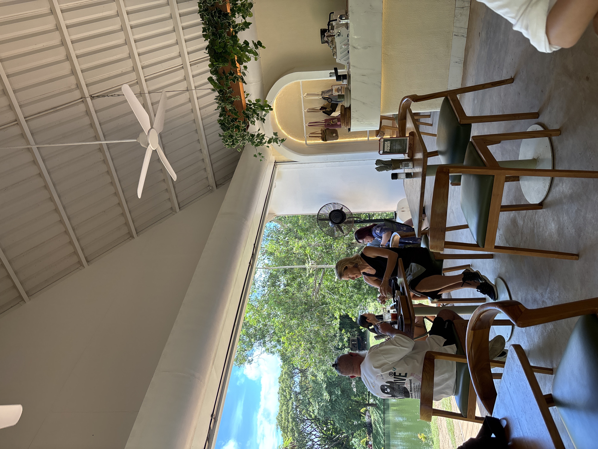 The image shows the interior of a restaurant or cafe, likely in Kuta Selatan, Indonesia. The building has a white, high-pitched roof with a visible ceiling fan. Green foliage is hung along the interior. Large windows offer a view of trees and bodies of water, with a visible fan in the window. The interior features wooden chairs and tables where people are seated. One woman is seated at the table, wearing a black tank top and shorts. A man in a white shirt with writing on the back is seated opposite her. The bar in the background shows coffee machines and purses.