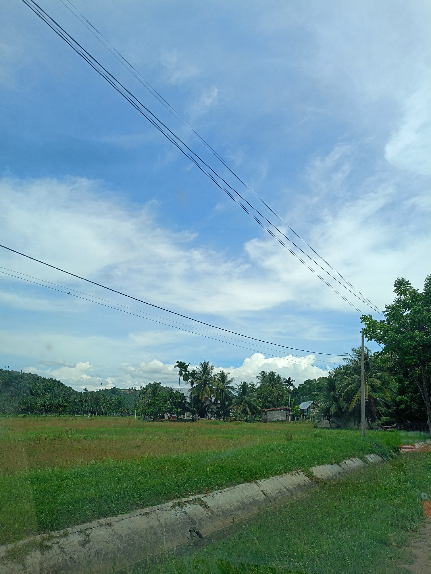 The image depicts a rural landscape in Seuneuboklhong, Indonesia. A field of mixed green and brown grass dominates the foreground, bordered by a concrete drainage ditch. Beyond the field, clusters of palm trees and other lush vegetation surround several simple, single-story buildings, suggesting a small settlement. A low, tree-covered hill rises in the background. Overhead, power lines stretch across a sky filled with scattered clouds and patches of blue. There are no visible people or animals.