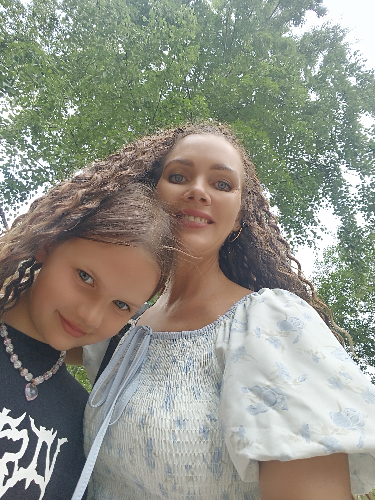 Two females, one adult and one child, are posing for a selfie outdoors. The adult female has long, curly brown hair and is wearing a light blue and white floral print top with puffed sleeves. She is smiling and looking at the camera. The child has brown hair with blonde highlights and blue eyes. She is wearing a black t-shirt with a white graphic design and a beaded necklace with a pink heart pendant. She is also smiling and looking at the camera. The background consists of lush green trees and a cloudy sky. The image appears to have been taken on a bright, overcast day. The specific location in Chaikovsky, Russia is not discernible from the image itself.