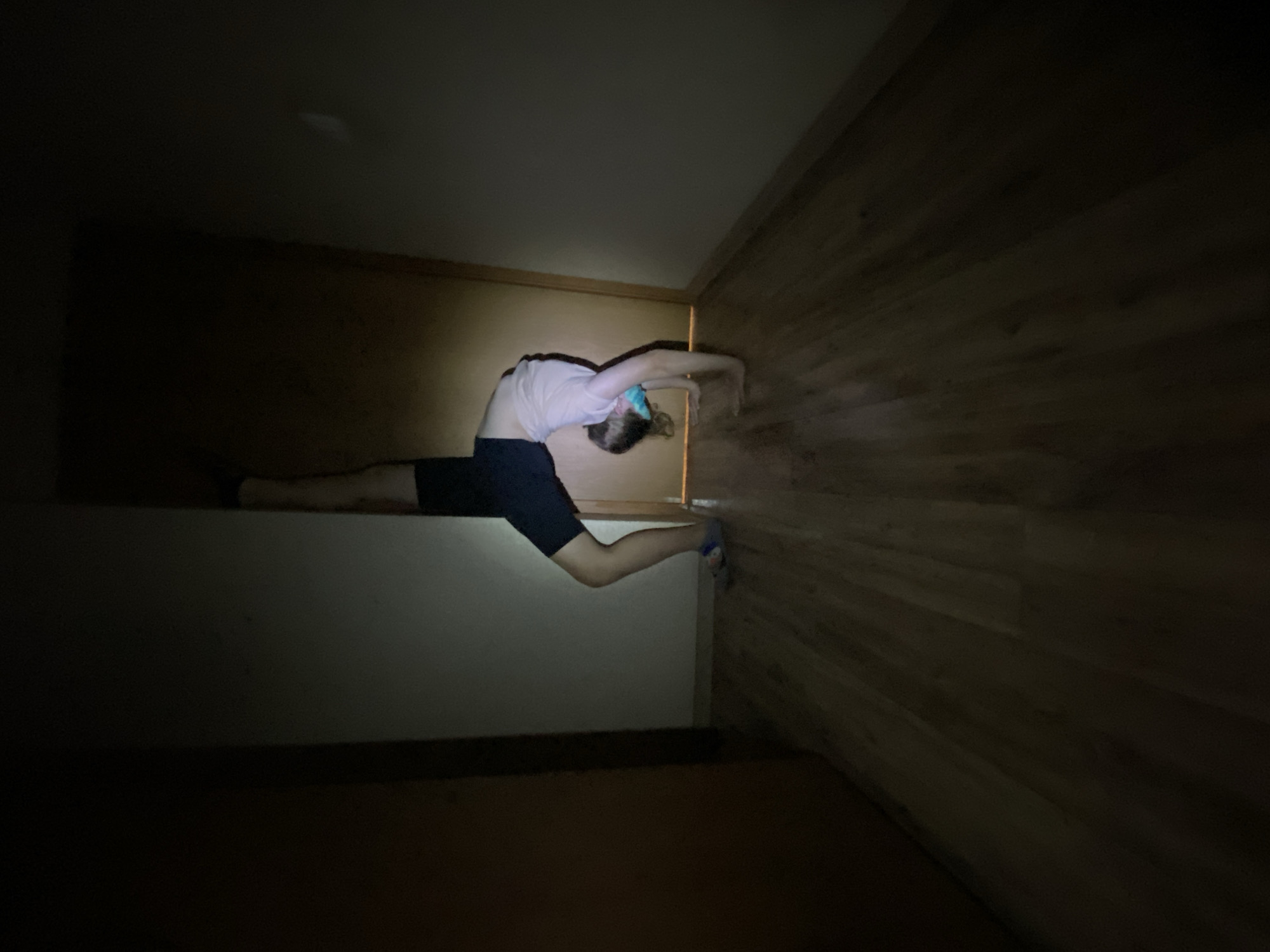 A dimly lit interior shows a person in a bridge position with a leg extended for a split. The person wears dark shorts and a light-colored top. Their hands touch a wooden wall. They are positioned between a wall and a surface. The environment is dark, obscuring details of the room.