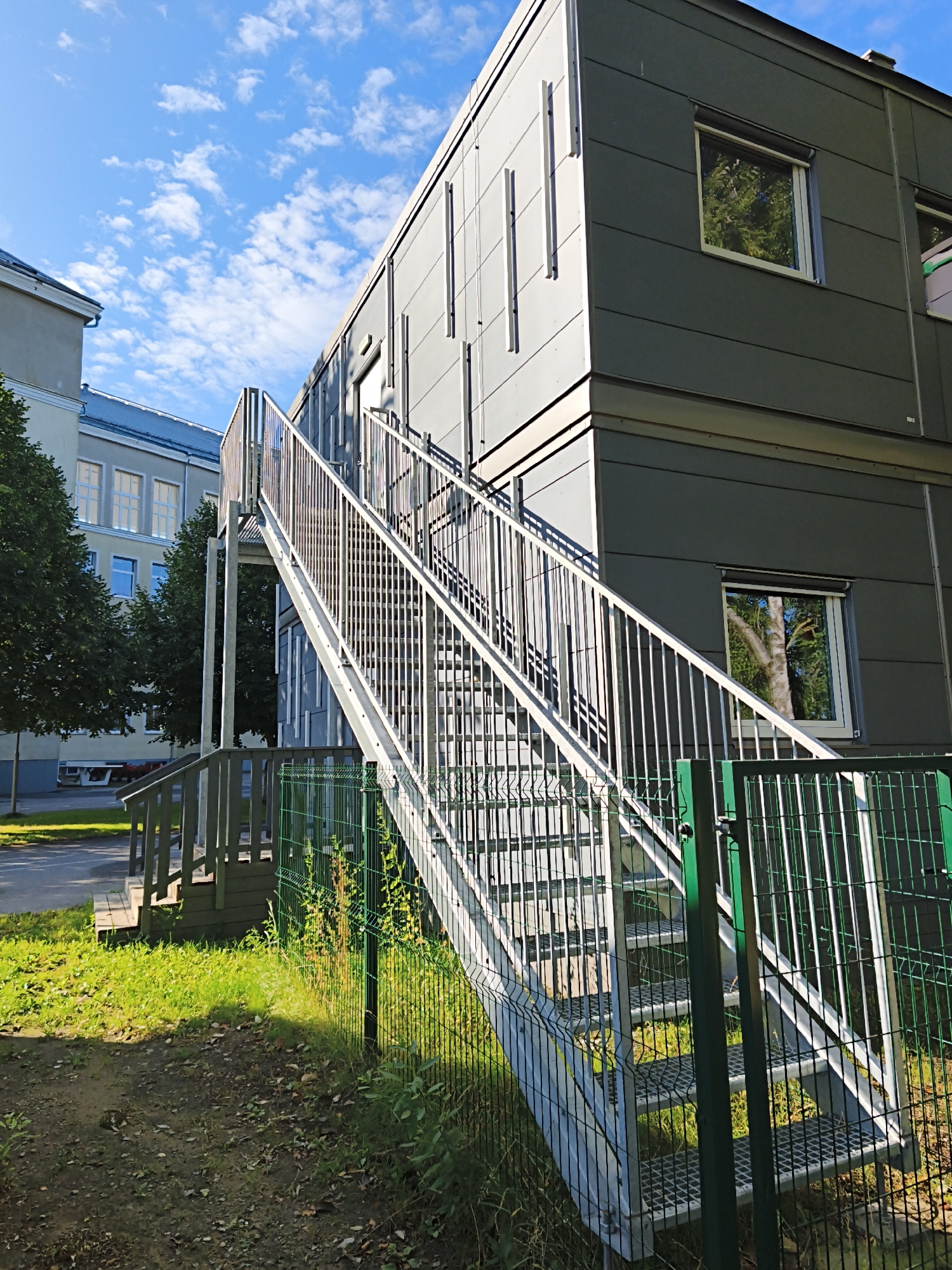 The image shows a modern, two-story modular building with a gray facade. An external metal staircase with railings leads up to the second story. Adjacent to the modular building is a lighter-colored, multi-story building with a sloped roof. Trees with green foliage are visible between the two buildings and to the left. In the foreground, a green wire mesh fence runs horizontally, separating the grassy area and a dirt path from the staircase and the buildings. The sky is blue with scattered white clouds. The scene appears to be an exterior view of a building complex, possibly a school or administrative facility. There are no people visible in the image. The architectural style and the presence of a modular building suggest a contemporary urban or suburban setting. While the image does not provide explicit clues to pinpoint Tallinn, Estonia, the question itself suggests this is the location.