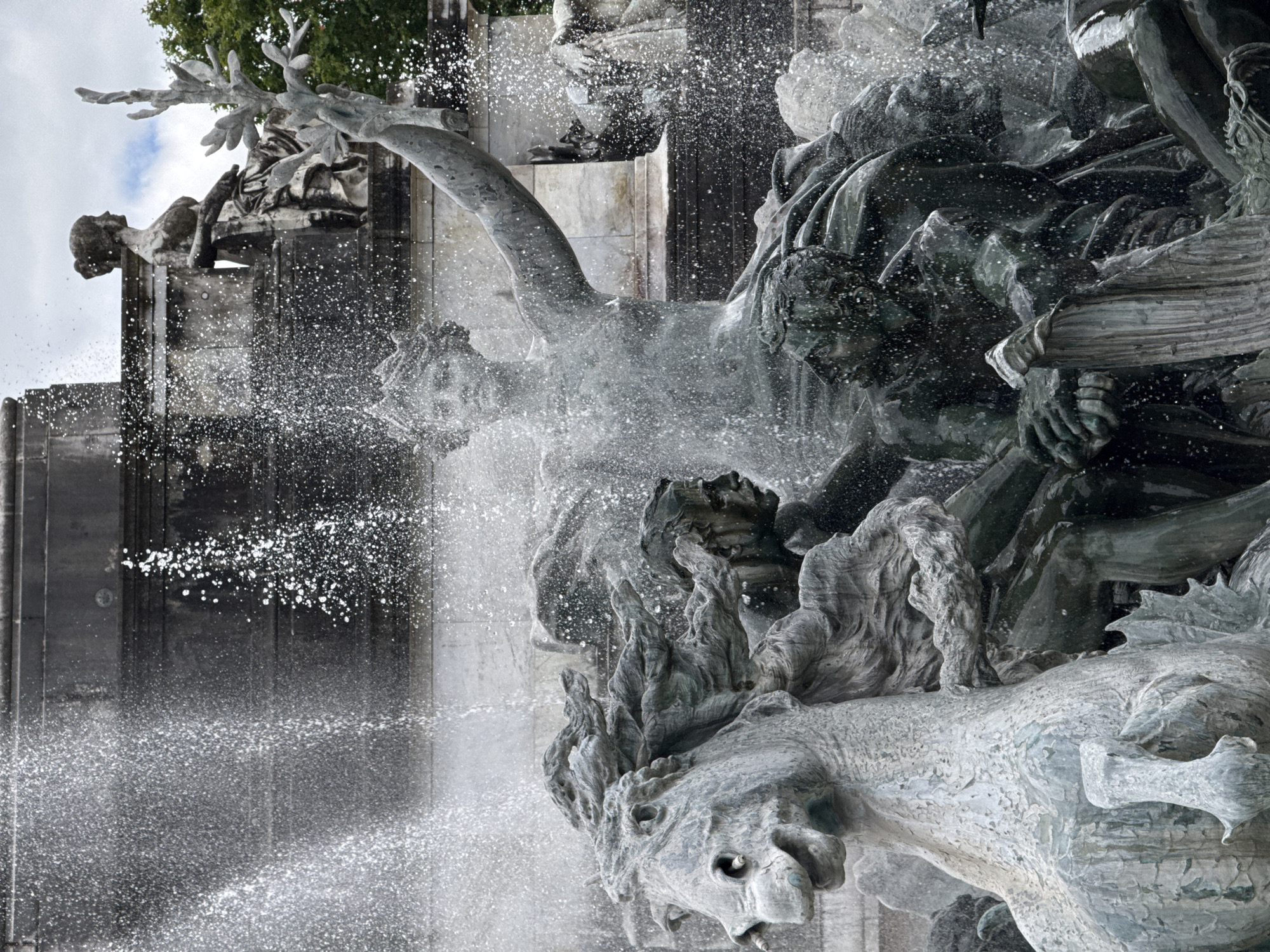 The image depicts the Fountain of the Girondins in Bordeaux, France. The fountain features a large central allegorical sculpture of Liberty trampling ignorance and despotism, surrounded by horses and human figures, all enveloped in cascading water. In the foreground, a stone horse's head with flowing mane is prominently visible, partially obscured by water spray. Behind it, several dark bronze human figures are depicted in dynamic poses, some seemingly struggling or in motion, with their faces and musculature detailed. Further back, a larger female statue, representing Liberty, is seated and holding a triumphant gesture. In the upper background, another female statue is perched, looking to the left. The fountain is set against a backdrop of grey stone architecture and trees, under a cloudy sky. The water from the fountain creates a dynamic and energetic atmosphere, with droplets catching the light and blurring the details of the sculptures.