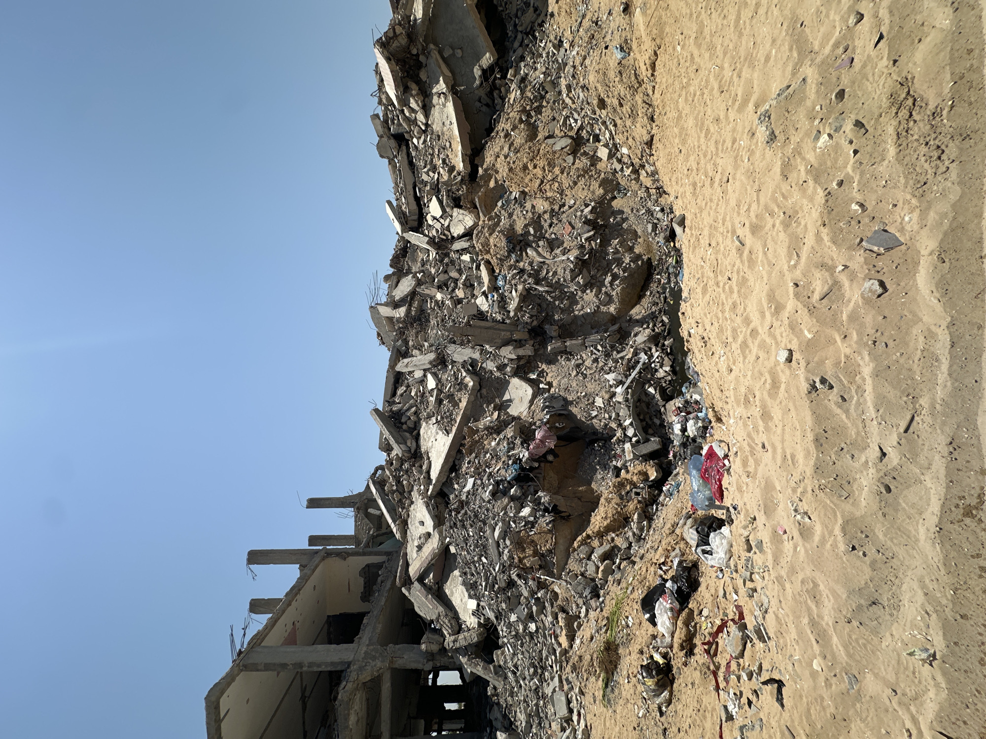 The image shows a large pile of rubble from a destroyed building in the city of Gaza, Palestinian Territories. The building appears to be made of concrete and rebar, and it is heavily damaged. There are pieces of concrete, broken walls, and twisted metal scattered throughout the rubble. Some personal belongings, such as clothing and plastic bags, are visible amongst the debris, suggesting the building may have been residential. The rubble pile is situated on sandy ground, with a clear blue sky overhead. The scene conveys a sense of destruction and abandonment.