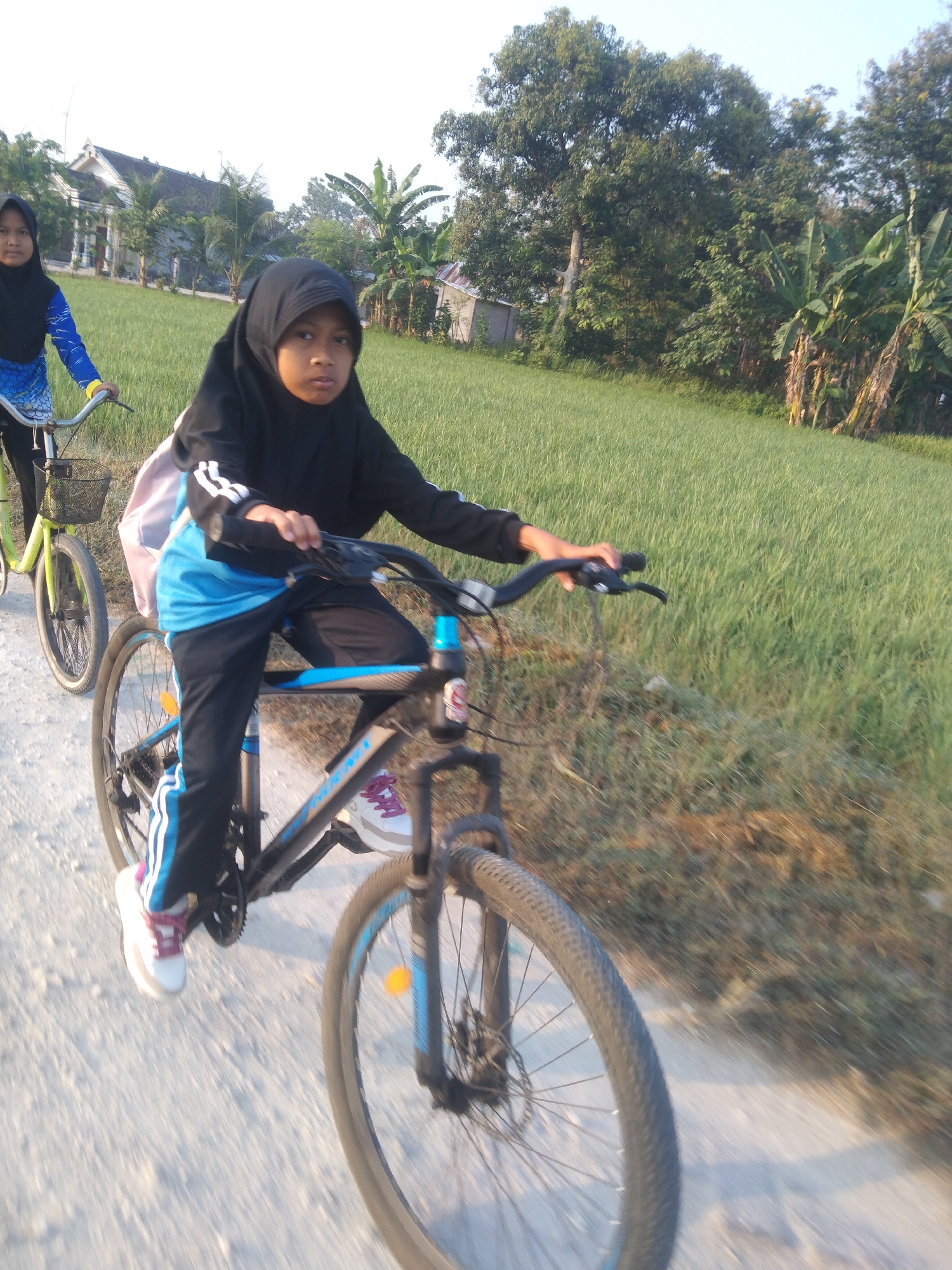 The image shows two young girls riding bicycles on a dirt road in Buntalan, Indonesia. The girl in the foreground, wearing a black hijab and a blue shirt, is looking at the camera while riding her bicycle. Her bicycle is black with blue accents. The girl in the background, also wearing a hijab, is riding a yellow bicycle and is further down the road. The environment is rural, with green fields and trees visible in the background. The sky is bright and appears to be early morning or late afternoon. The road surface is dirt and gravel.