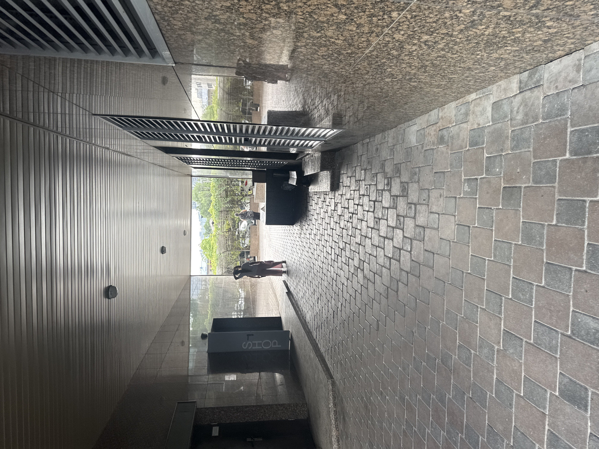 The image depicts a covered walkway with a stone-paved floor. Two individuals are visible in the distance. One person is standing near a structure with the word "SHOP" written vertically on it. Another person is further down the walkway, with their back to the camera. The walkway is flanked by walls, one of which has a granite-like texture, and features vertical ventilation grates. The ceiling of the walkway has a ribbed metallic appearance. Trees and buildings are visible beyond the walkway's opening. The scene suggests an urban environment, possibly a commercial or public area in Almaty, Kazakhstan, given the context.