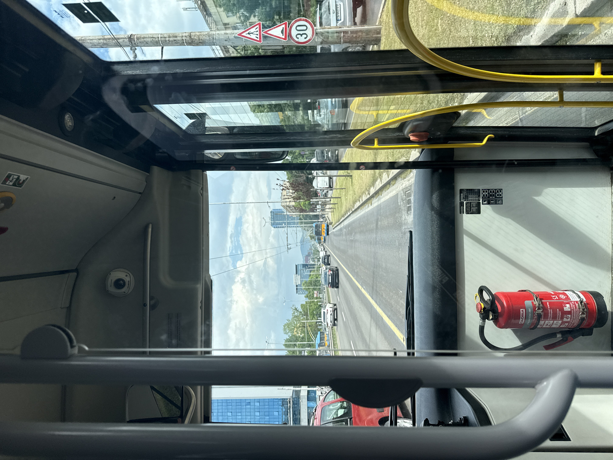 The photo, taken from inside a bus, shows a view of a street in Sofia, Bulgaria. The street has multiple lanes, with cars and buses traveling on it. A yellow line marks the lane dividers. Tall buildings are visible in the background. Inside the bus, a red fire extinguisher is mounted on the wall. A yellow grab rail is visible above. A speed limit sign (30) is seen through the bus window.