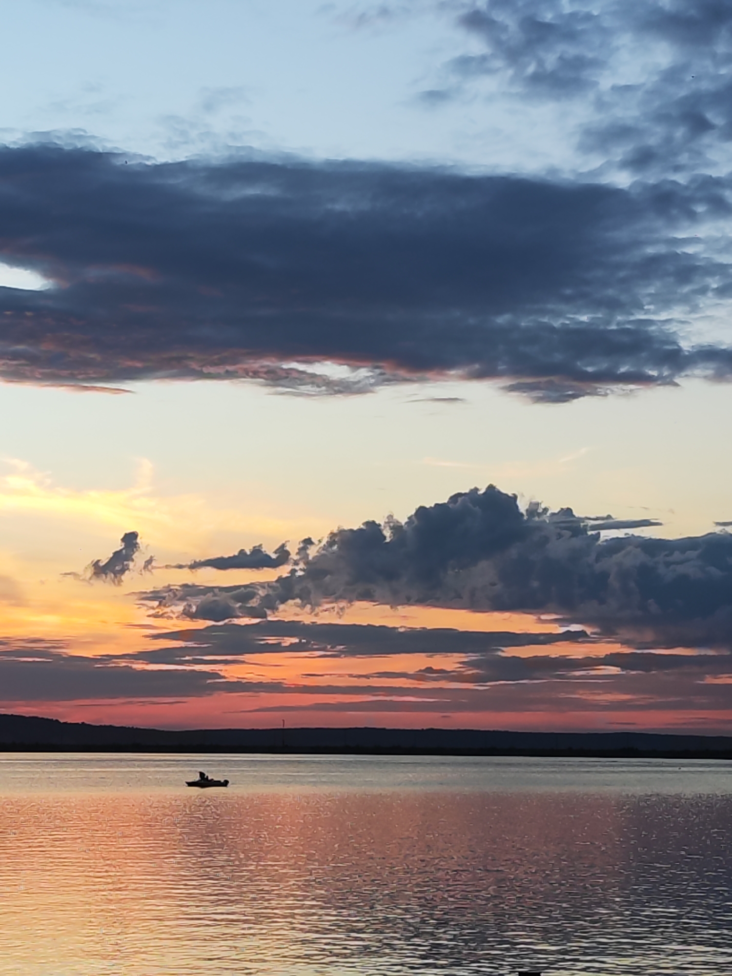 The image depicts a serene sunset over a wide body of water, likely a river or lake, near Chaikovsky, Russia. The sky is a canvas of dramatic colors, transitioning from a pale blue at the top to vibrant oranges and pinks near the horizon, interspersed with dark, brooding clouds. These clouds are textured and varied, with some appearing wispy and others more dense and imposing.
In the distance, a dark silhouette of the landmass forms a horizon line, suggesting a natural or rural setting. A single, small boat with at least one person is visible on the water, appearing as a tiny dark speck against the vast expanse. The water itself reflects the fiery hues of the sunset, creating shimmering patterns of orange, pink, and purple across its surface.
The time of day is clearly sunset, indicated by the warm, low-angled light and the colors in the sky. The weather appears partly cloudy, contributing to the dramatic visual display. The overall mood of the image is one of tranquility and natural beauty, with the solitary boat adding a subtle element of human presence within the grand landscape. There is no visible text in the image.