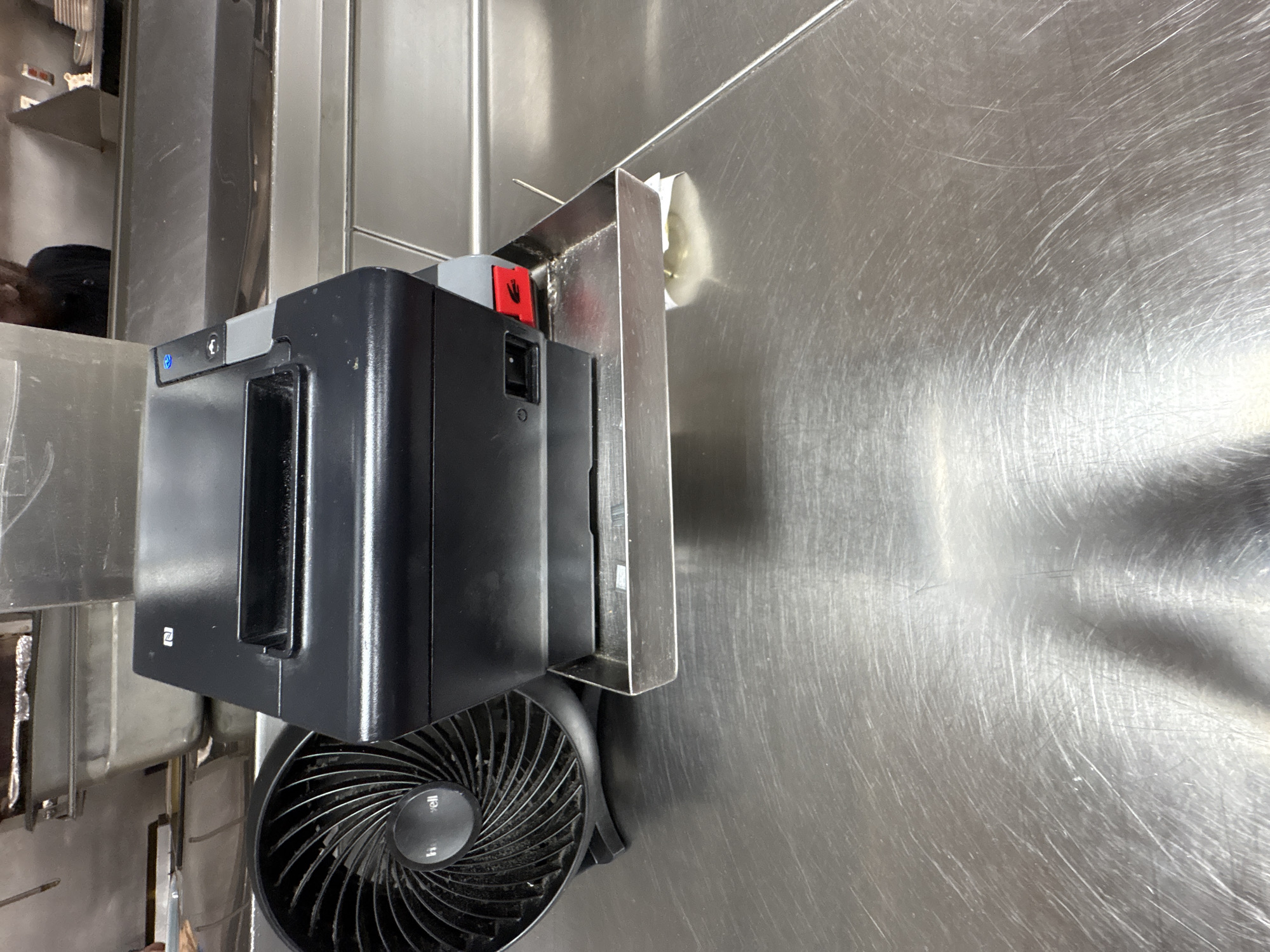 This media file captures a close-up view of a black electronic device, possibly a printer or scanner, mounted on a stainless steel surface. To the left of the device, a partially visible shelf holds what appear to be cleaning supplies. A person wearing a dark beanie is visible in the background, peeking over the shelf. Below the electronic device, a black fan with a circular grille is positioned on the stainless steel surface. The fan has a central circular emblem with text that is partially legible, appearing to read "Honeywell". The stainless steel surface exhibits numerous scratches, indicating wear and tear. The lighting suggests an indoor setting, possibly a commercial kitchen or utility area. There is a red lever with a white hand icon on the side of the electronic device, indicating a safety or operational feature. The scene appears to be set in Panama City Beach, USA. No specific activity is depicted, and the overall impression is one of a functional, everyday environment.