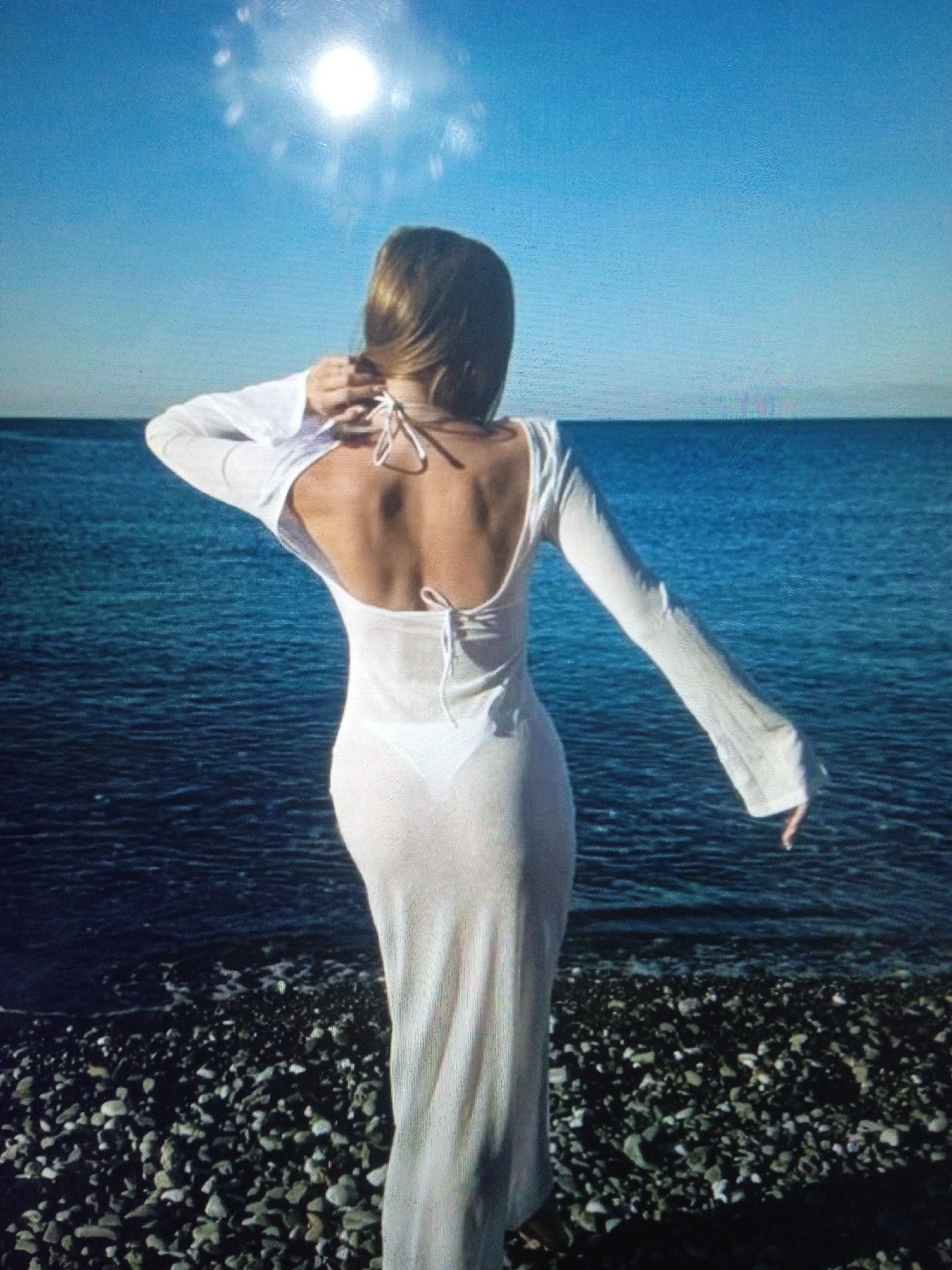 The image depicts a woman standing on a rocky shore, facing away from the camera. She is wearing a sheer, white, long-sleeved dress over a white bikini. Her hair is light brown and is partially pulled back. The ocean is visible behind her, with calm water under a bright blue sky. The sun is high in the sky, creating a bright glare. The scene appears to be a beach. The location is not specified as Chernivtsi, Ukraine.