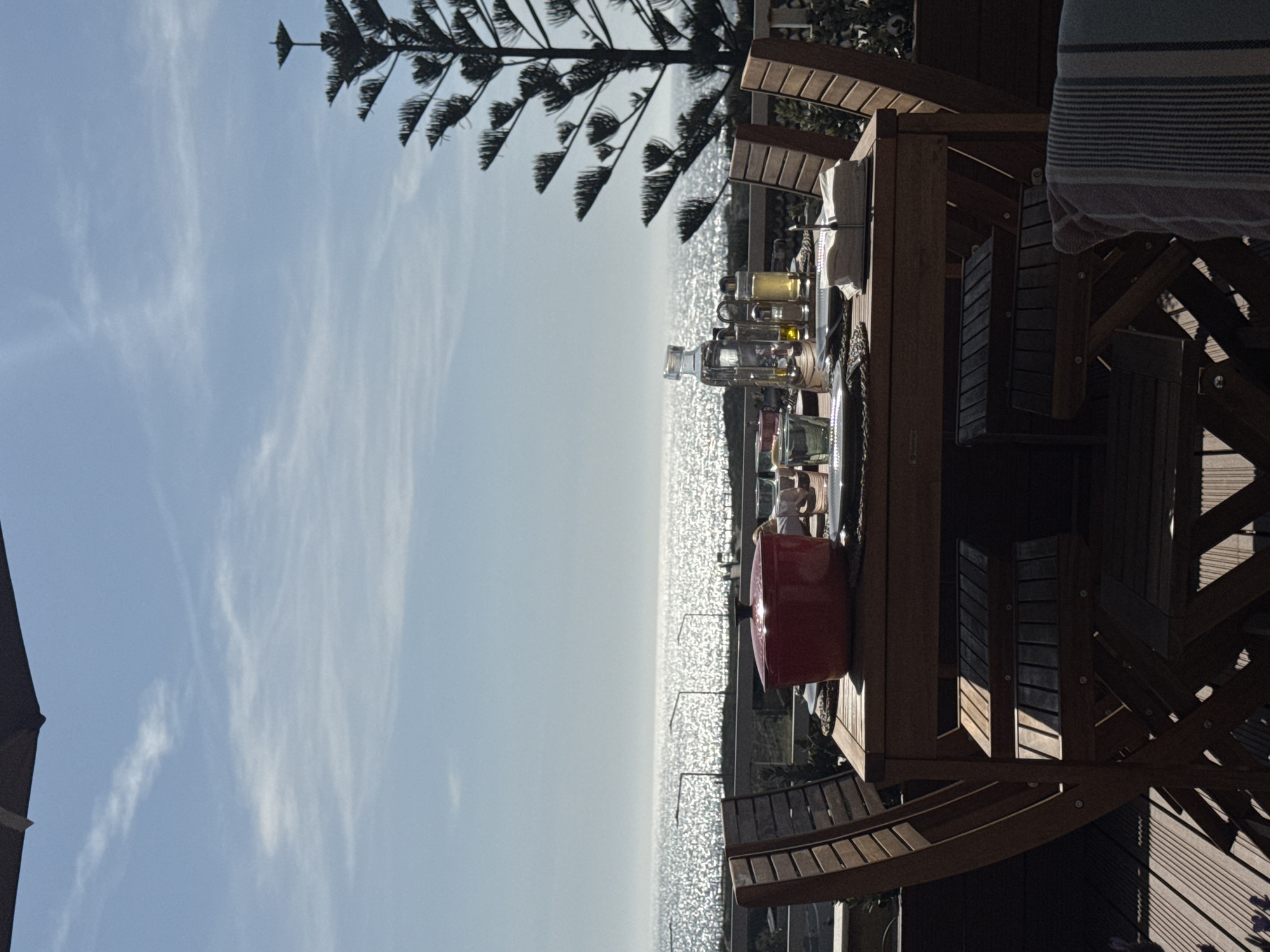 The image depicts an outdoor setting on a sunny day, likely a patio or deck, overlooking a vast expanse of sparkling water, presumably the ocean or sea, in Vila Nova de Gaia, Portugal. The image is oriented vertically but should be viewed as a horizontal landscape.
In the foreground, a sturdy wooden table and chairs are set on a wooden deck. On the table, there are several glass bottles containing liquids of varying colors (clear, yellow, amber), suggesting beverages, oils, or condiments. A prominent red lidded pot or casserole dish sits centrally on the table. A striped cloth, predominantly blue and white, is draped over a chair or railing on the far right.
To the left, a tall conifer tree with distinctive, layered branches (possibly a Norfolk Island Pine) extends into the sky, framing the scene. The background is dominated by the shimmering water, intensely illuminated by sunlight, creating numerous bright reflections across its surface. A clear horizon separates the water from the sky. The sky above is a bright blue with some sparse, wispy white clouds. Part of a dark umbrella or shade structure is visible on the far left.
No people are visible in the scene. The arrangement of items on the table suggests a setup for an outdoor meal or gathering. The bright sunlight, clear sky, and sparkling water indicate good weather, likely late morning or early afternoon. The overall atmosphere is one of peaceful outdoor enjoyment with a beautiful scenic view. No visible text is discernible in the image.