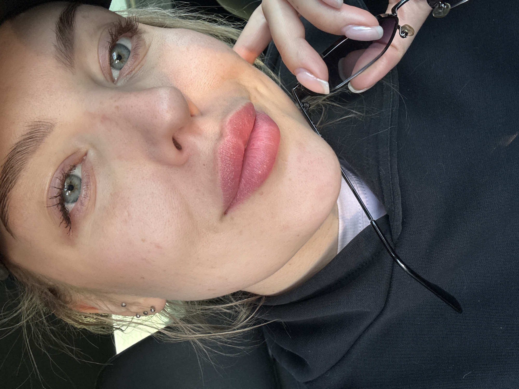 A close-up, angled selfie captures a light-skinned woman with fair hair and light-colored eyes. She is holding black-rimmed eyeglasses in her right hand, with the fingers displaying long, manicured nails. Her lips are notably defined and colored, while her skin shows slight imperfections. She wears a black hooded sweatshirt over a light-colored shirt. The background is blurry, but the scene is set outside, likely in daytime with natural lighting. Multiple earrings are visible in her left ear. There is no visible indication that this image was taken in Sherlovaya Gora, Russia.