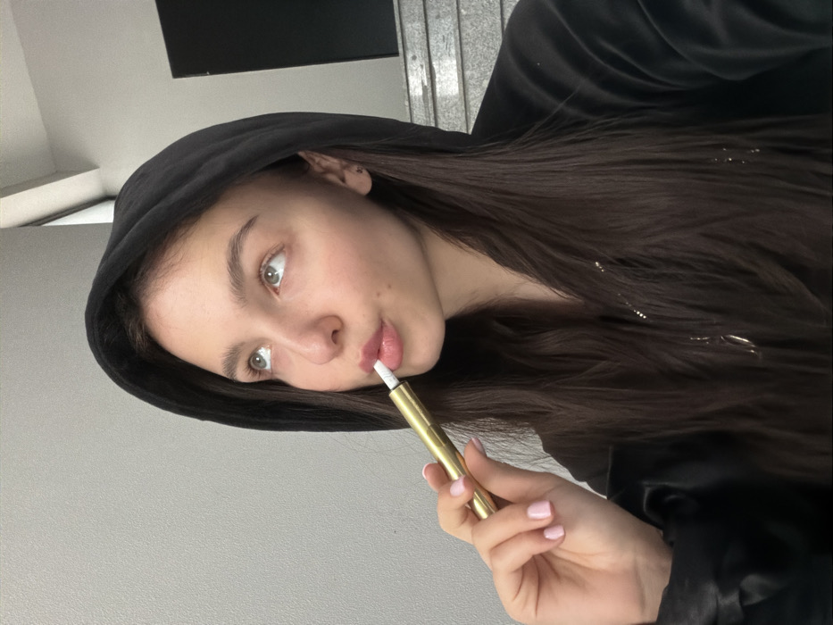 A young female is depicted in a medium close-up, looking upwards and slightly to her left. She has long, dark brown hair and is wearing a black hooded garment, consistent with velvet or velour material. Her right hand, adorned with light pink nail polish, holds a golden, cylindrical device, identified as a vape pen, to her slightly pursed lips. A white tip from the device is positioned within her mouth. Her eyes are light-colored, and she wears small, dark stud earrings. The background features a plain, light grey wall on the left, transitioning to a dark-colored, multi-tiered structure resembling stairs on the right. No visual identifiers are present within the image to confirm a specific location within Sofia, Bulgaria.