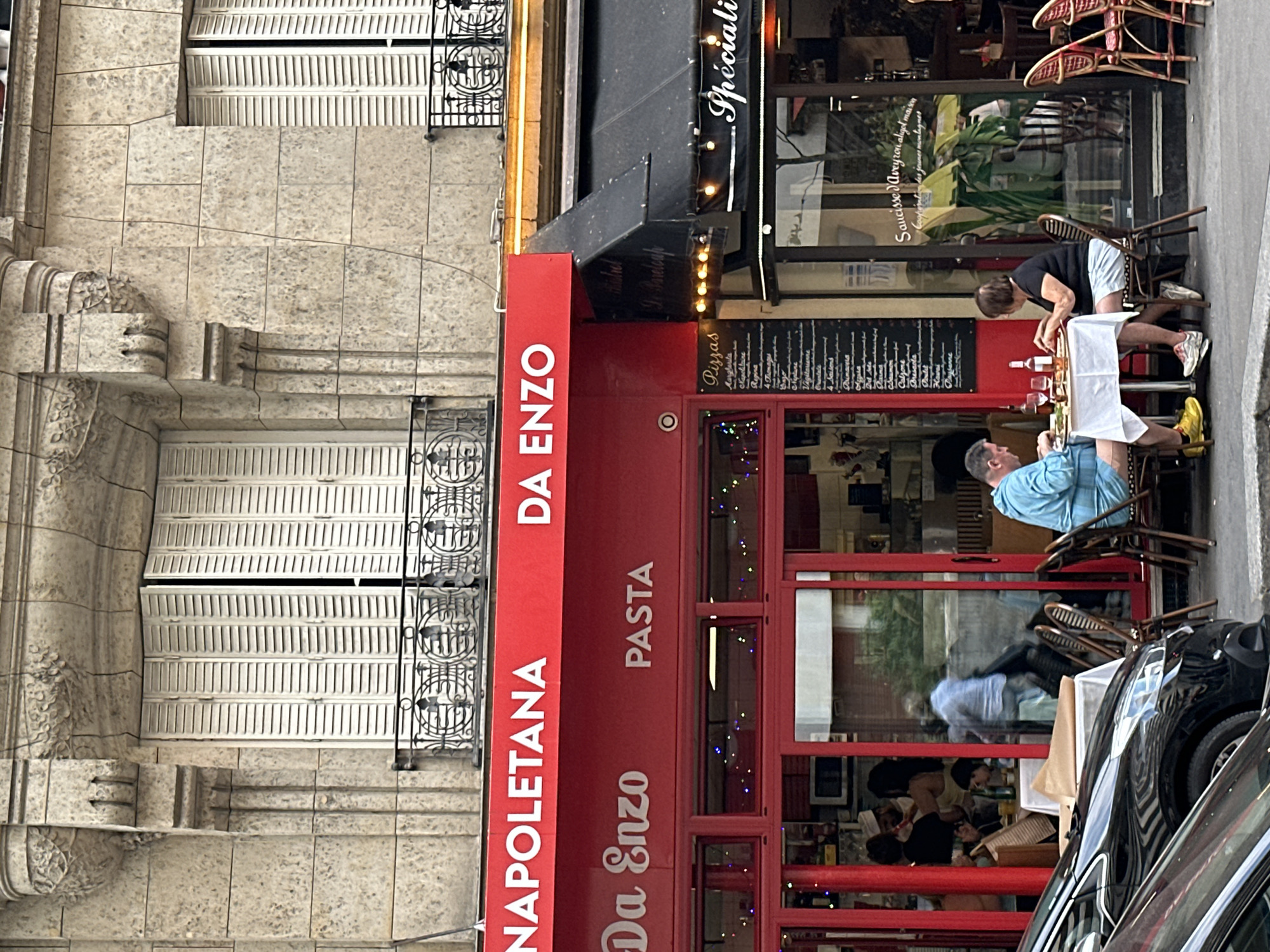 This is a street-level photograph of a restaurant scene in Paris, France. The facade of the restaurant, "Da Enzo NAPOLETANA PASTA", is prominently displayed in red with white lettering. Several patrons are seated at tables outside the restaurant, eating and drinking. In the foreground, two individuals, a man in a blue shirt and shorts and a woman in a black shirt and shorts, are seated at a white-clothed table, interacting with their food. To the right of the restaurant, another establishment with a black awning is visible, also with outdoor seating. Cars are parked on the street in the lower left quadrant of the image. The buildings in the background feature stone facades and white shutters, characteristic of Parisian architecture. The overall impression is of a lively, casual dining environment on a city street.