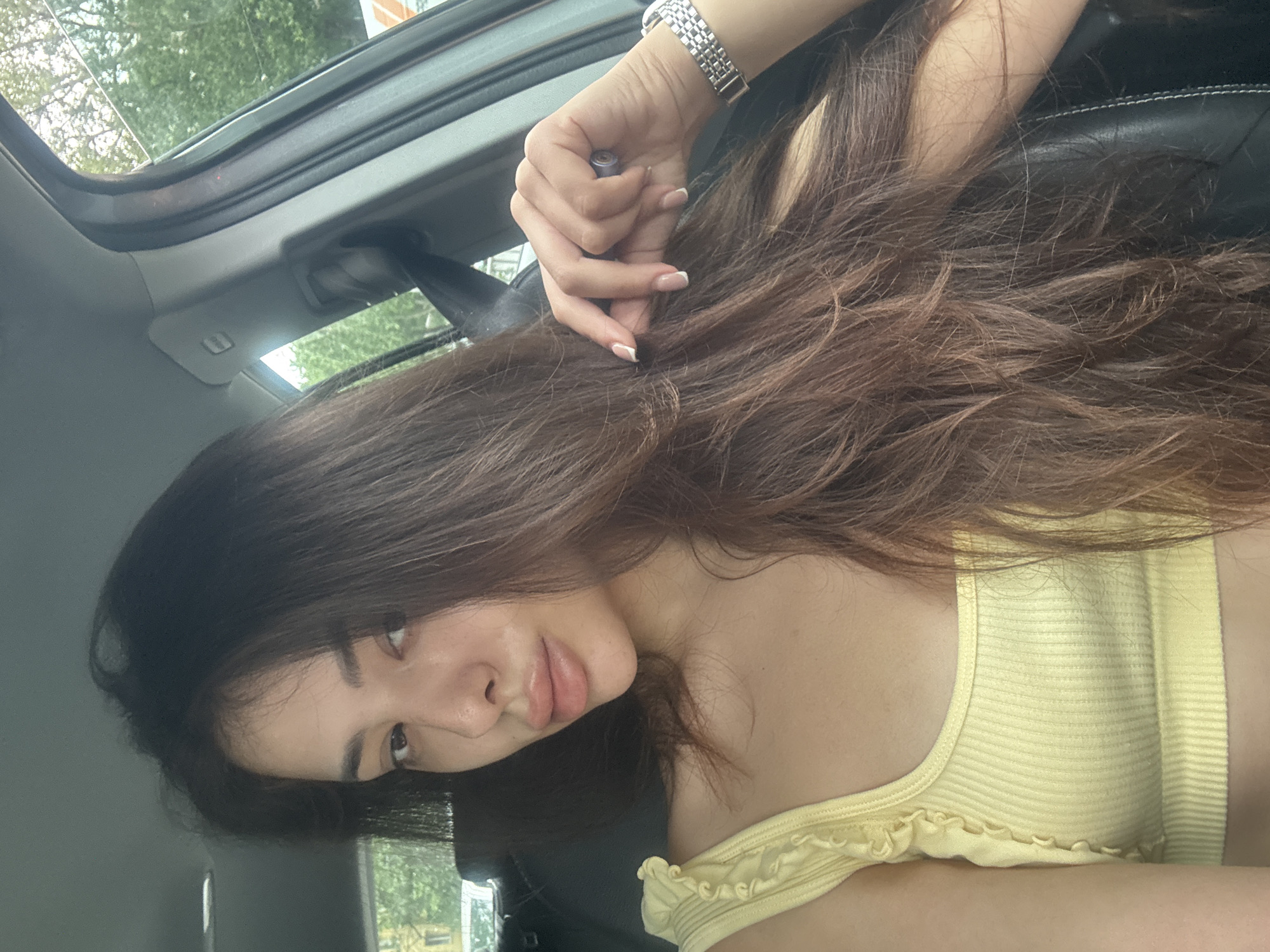 The image displays a young woman inside a car, rotated 90 degrees counter-clockwise. She has long, wavy dark brown hair that covers much of the frame. She is wearing a light yellow ribbed tank top with ruffled detailing along the neckline and straps. On her right wrist, she wears a silver-toned metal wristwatch, and her nails feature a French manicure with white tips. Her right hand, holding a small dark cylindrical object (possibly a vape pen), is raised and rests near her hair.
The woman has fair skin and a neutral, slightly pouting expression, looking directly at the camera. Her eyes are dark.
The setting is the interior of a car. Visible elements include a gray headliner, a sunroof through which green foliage and parts of the sky are visible, and glimpses of dark car seats. Natural daylight illuminates the scene, suggesting it is daytime. The soft lighting indicates either an overcast day or dappled light filtering through trees. The overall activity appears to be posing for a casual photo or selfie within the car. There is no visible text within the image. The location context provided is Plovdiv, Bulgaria.