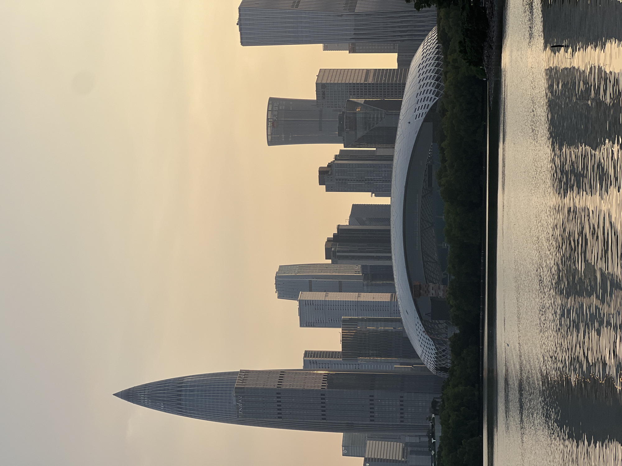The image shows a waterfront view of the Shenzhen Bay Park in Nanshan District, China. In the background, a modern cityscape is visible with numerous skyscrapers of varying heights and architectural styles. One prominent skyscraper, the Shenzhen Ping An Finance Centre, stands out with its distinctive pointed top. In the foreground, a body of water reflects the golden hues of the sky, suggesting either sunrise or sunset. A thin, white bird is seen standing on the water. A large, curved dome-shaped structure, possibly a stadium or convention center, is situated between the water and the skyscrapers, partially obscured by a line of dense green trees. The sky is a soft, hazy gradient of orange and yellow.