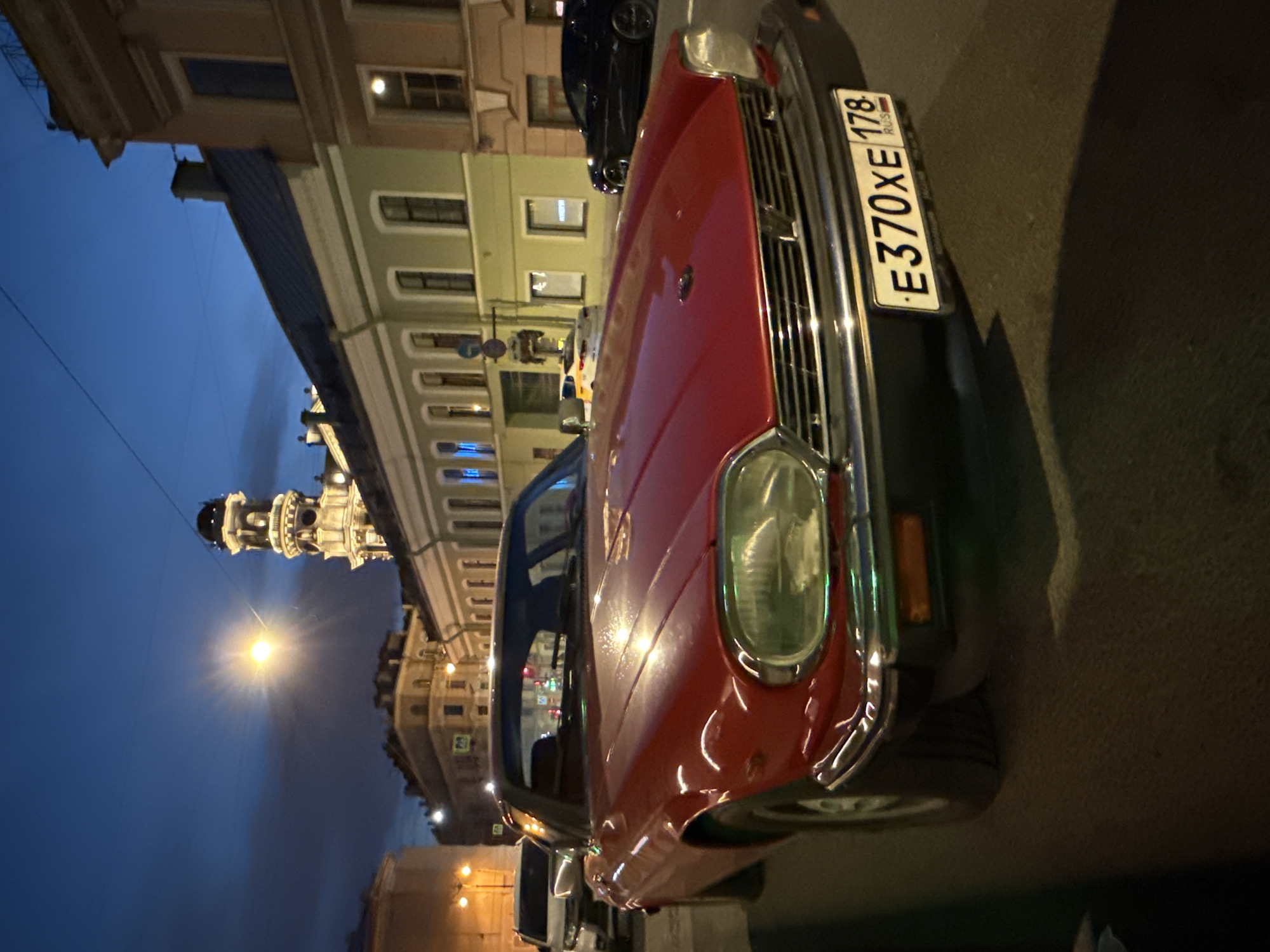 An eye-level, medium shot captures a red classic Jaguar XJ-S parked on a street in Saint Petersburg, Russia, at twilight. The car is positioned at an angle, with its front end slightly to the right and its headlights illuminated. The car's license plate is clearly visible, reading "E370XE 178 RUS". In the background, the historic buildings of Saint Petersburg line the street. A prominent clock tower, believed to be the Admiralty Spire, stands tall against the deep blue sky, illuminated by streetlights and the setting sun's glow. The scene is dimly lit, with streetlights casting a warm glow on the car and the surrounding architecture. No people are visible in the frame. The street appears to be paved with asphalt.