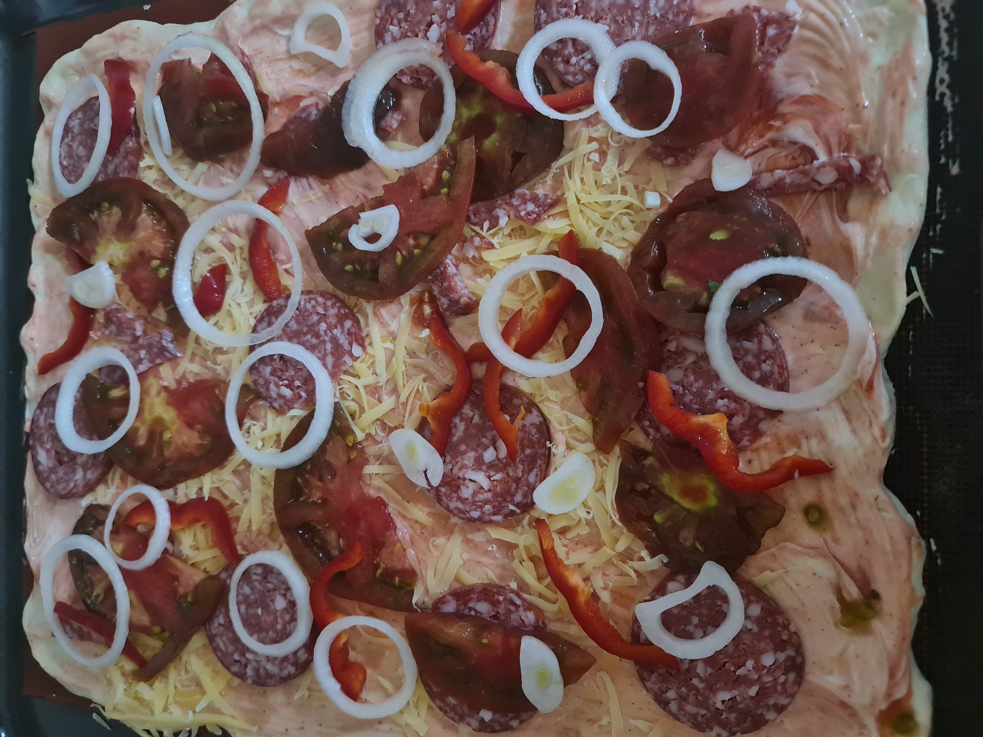 The image shows a close-up, overhead view of an uncooked pizza on a dark baking sheet. The pizza has a pinkish-red sauce base spread thinly over a light-colored dough. It is topped with shredded yellow cheese, slices of salami, sliced tomatoes, thin slices of red bell pepper, and rings of white onion. A few small pieces of garlic are also visible. The pizza is not in a specific location in Angarsk, Russia, as the image only depicts the food item itself.