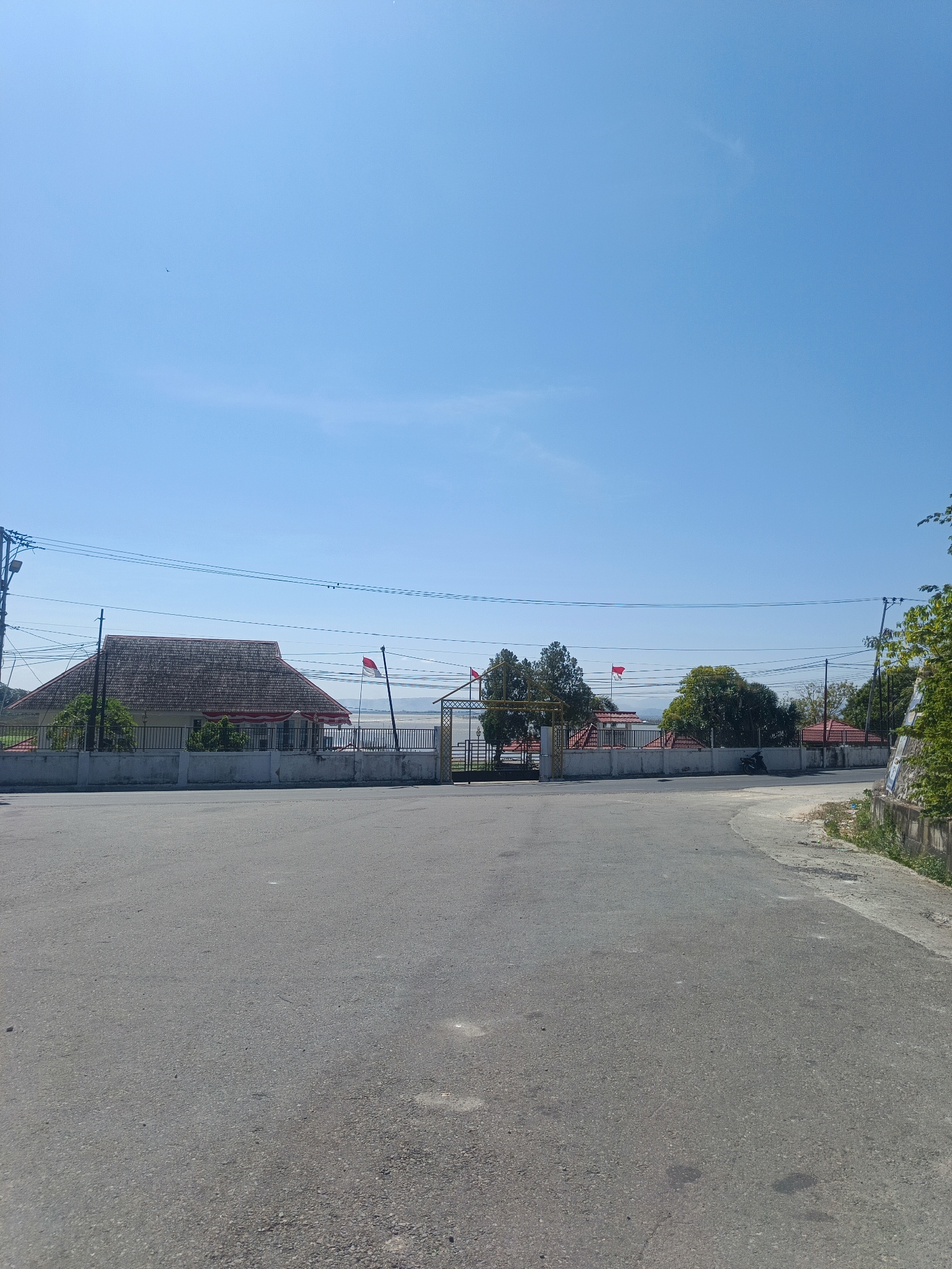 A paved road leads towards a fenced compound under a clear blue sky. The compound contains several buildings with red roofs and a large, gabled building with a thatched roof. Indonesian flags are visible on poles within the compound. A lone motorcycle is parked outside the fence. The environment is sunny and appears to be a rural or semi-rural area. The specific location in Ilotidea, Indonesia, is not discernible from the image.