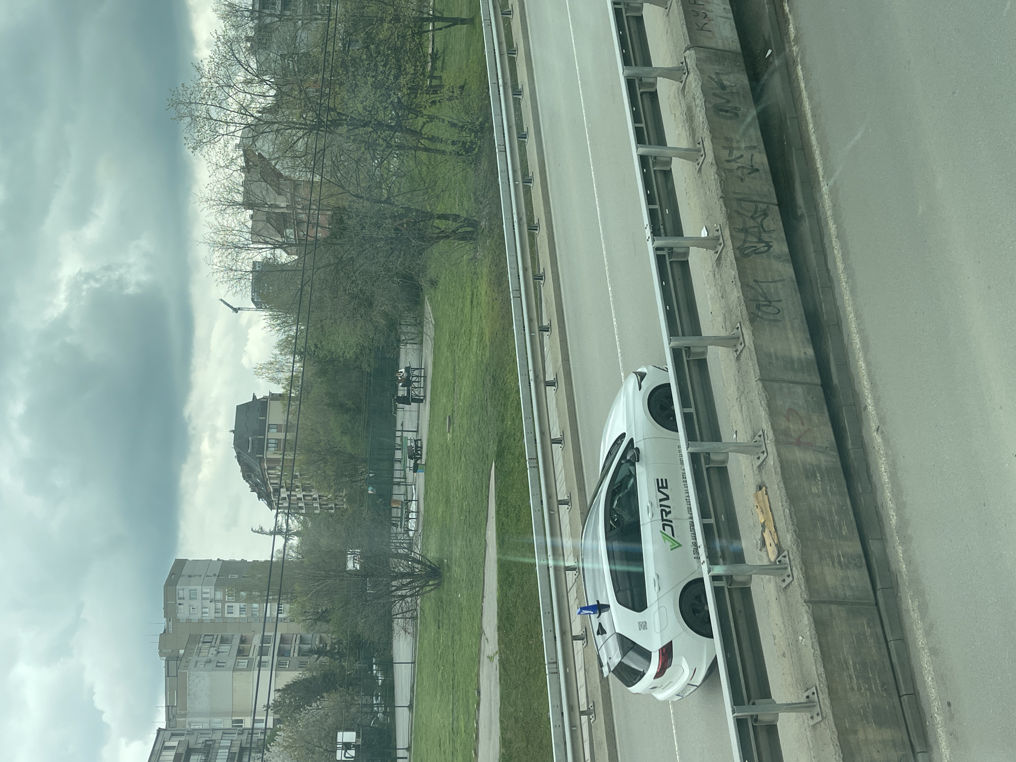The image shows a view from an elevated position, likely a bridge or building, overlooking a street and a park area in Sofia, Bulgaria. A white car with "V DRIVE" branding is parked along the roadside, next to a concrete barrier with graffiti. The street has multiple lanes and divides the green park space from the urban area with apartment buildings in the background. The sky is overcast.