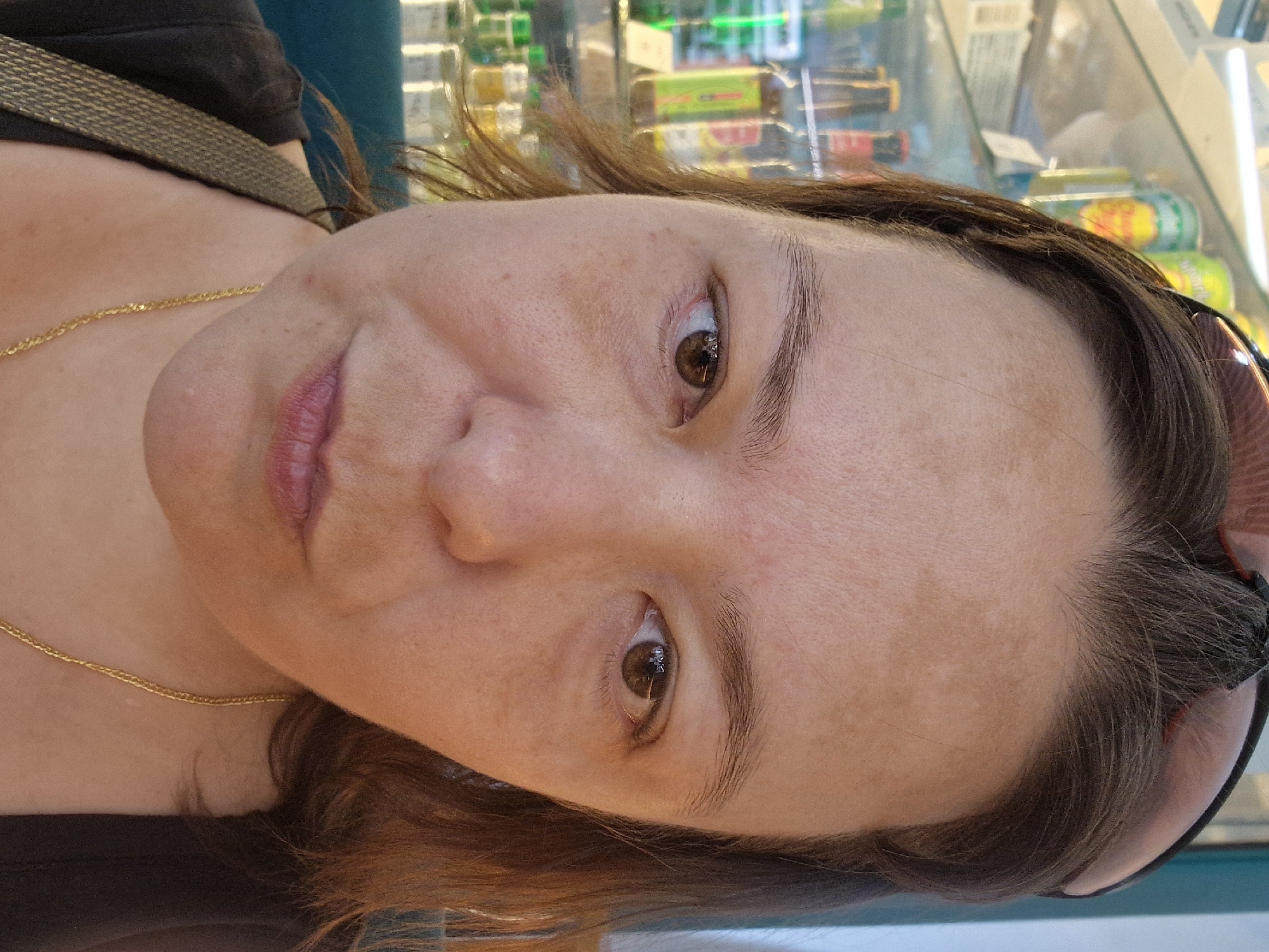 A close-up, frontal, eye-level selfie shows the upper body of a woman with light brown hair and brown eyes. She is wearing a dark top and a gold chain necklace. Her face has freckles and some sun spots. She is wearing sunglasses on top of her head. The background appears to be a store with shelves of products, possibly drinks in bottles and cans. The lighting suggests it is daytime and possibly indoors. The location is described as Unknown, Unknown.