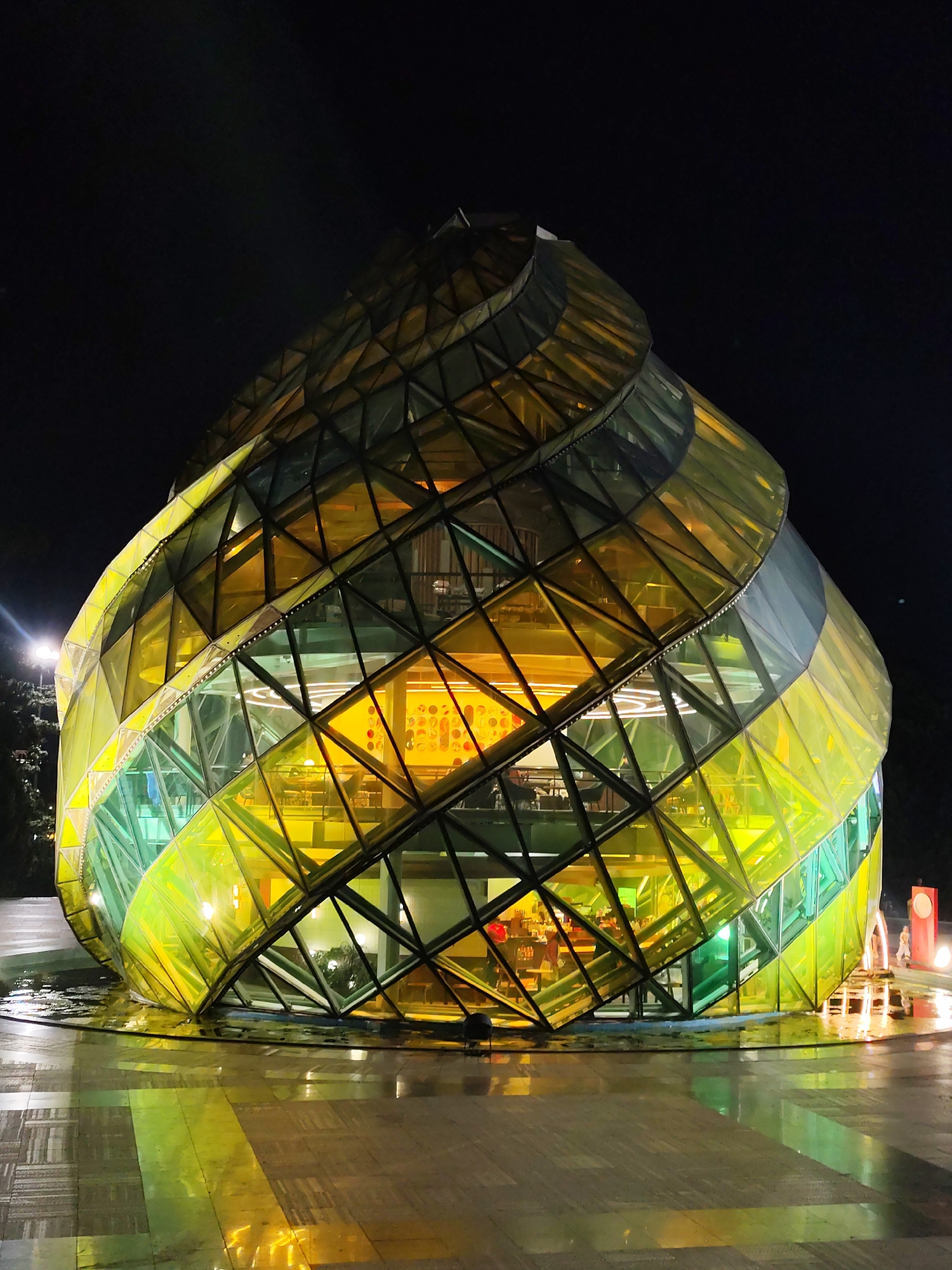 The image shows the stunning architecture of the Crazy House (Hằng Nga Villa) in Da Lat, Vietnam, at night. The building, designed by architect Đặng Việt Nga, features an unusual, organic shape reminiscent of a giant tree or a fantastical creature. Its exterior is composed of a complex lattice of metal and colorful, translucent glass panels, predominantly in shades of yellow and green, which are illuminated from within, creating a vibrant and otherworldly glow.
The structure is multi-leveled, with various interconnected rooms and walkways visible through the glass. The faceted glass creates a kaleidoscope effect, reflecting the internal lighting and the surrounding environment. In front of the building, the ground is wet and reflects the building's light, indicating recent rain or a fountain feature. A pathway of patterned paving stones leads towards the entrance. In the background, there are glimpses of trees and distant lights, suggesting the building is situated within a landscaped area of the city. On the right side of the image, a few figures are visible near a red, cylindrical structure and a light pole, possibly visitors or staff. The overall impression is one of a surreal and enchanting architectural masterpiece illuminated against the dark night sky.