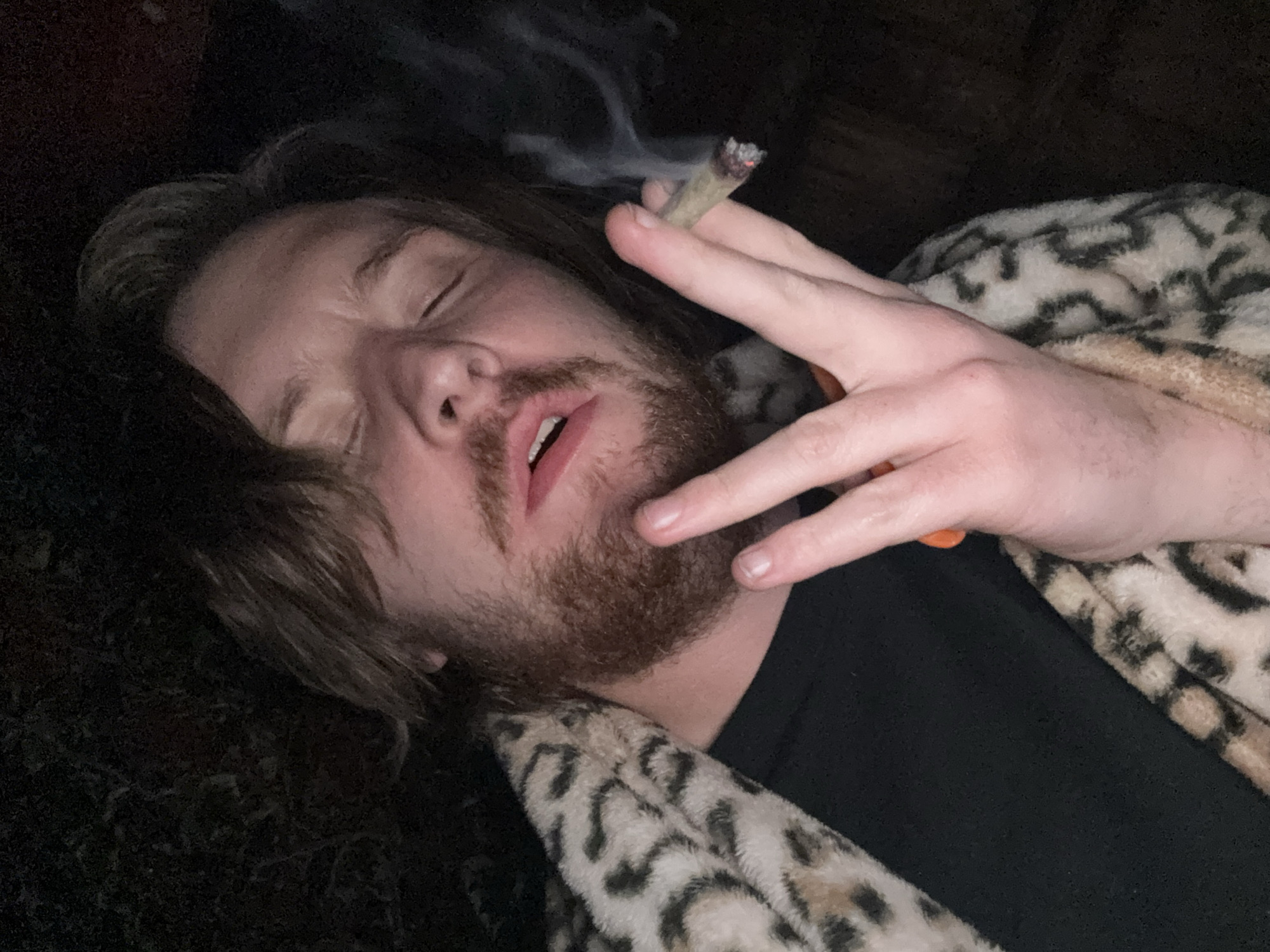 A person is reclining, likely in an indoor environment due to the low lighting and presence of soft furnishings. The individual has fair skin, light brown hair, a beard, and is wearing what appears to be a leopard-print robe or blanket over a black shirt. The person has their eyes closed and appears to be holding a lit cigarette or joint, with smoke emanating from the burning end. The background is dark and indistinct. The image does not contain any information to specify the location of the city of Cañada de Madero, Mexico.