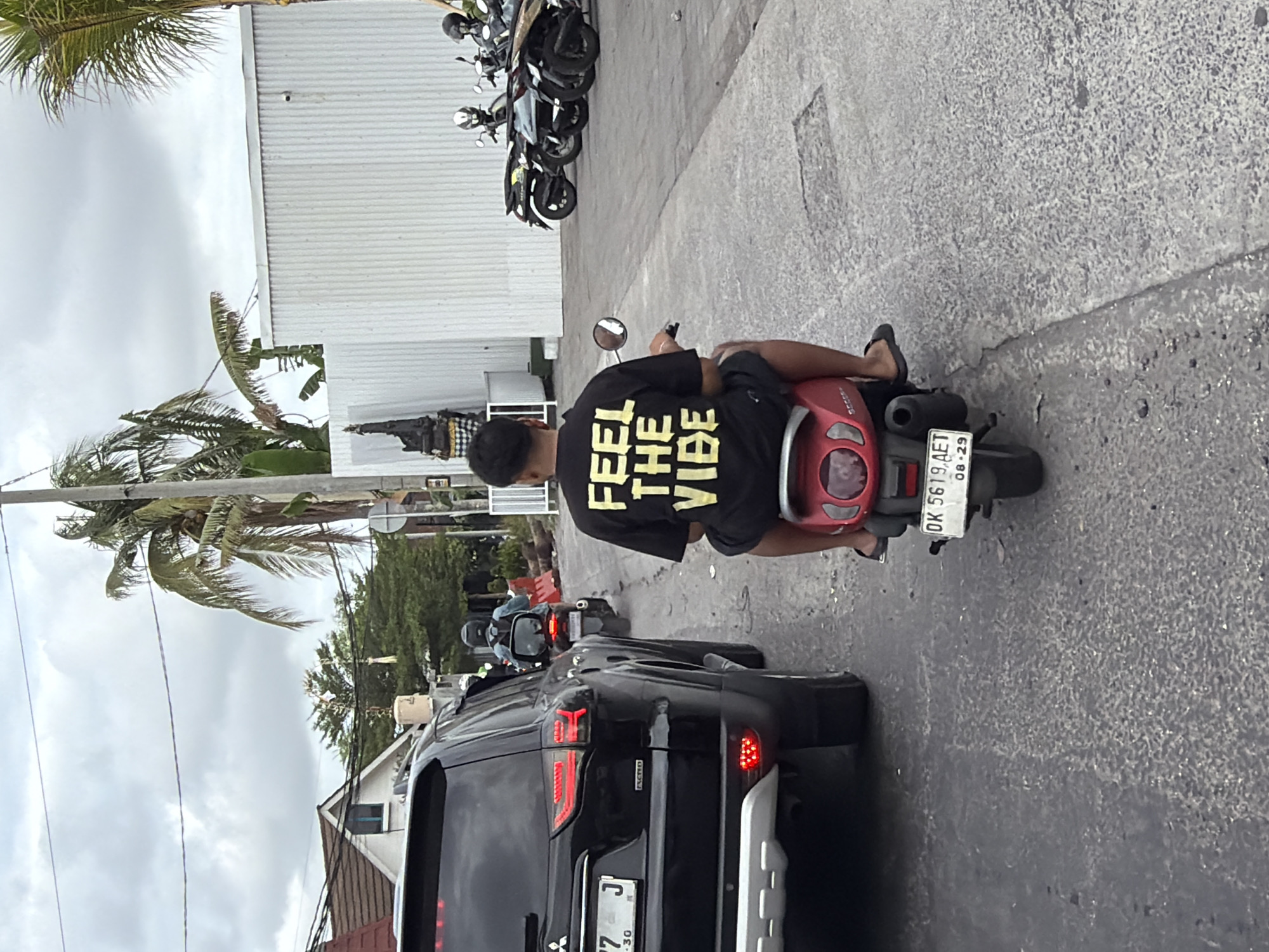 In North Kuta, Indonesia, on a cloudy day, a person sits on a red scooter, facing away from the camera. They are wearing a black t-shirt with yellow lettering that reads "FEEL THE VIBE" and black shorts. The scooter has a white license plate that reads "DK 5619 AET 08-29". A black SUV is in the foreground to the left, and other scooters and a few people on motorbikes are visible in the background. The scene appears to be on a paved road, with some buildings and vegetation visible on either side.