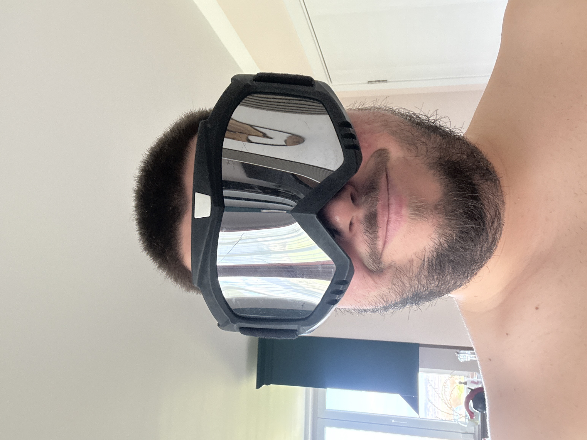 A man with dark hair and a beard is wearing black goggles. He is indoors, and a window is visible in the background. The location is not discernible from the image.