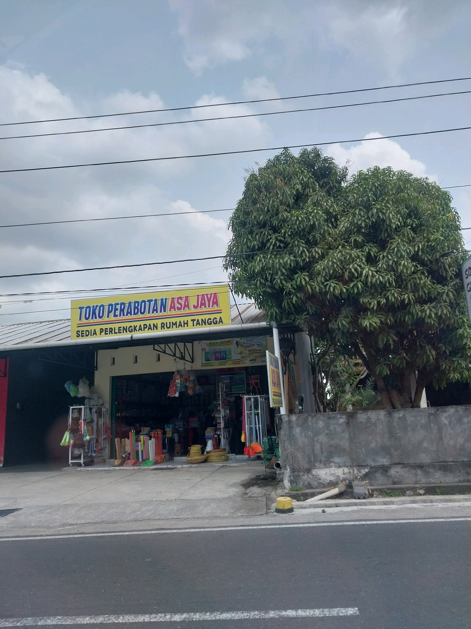 The image shows a street view of a small business in Tirtomartani, Indonesia. The business, identified by a yellow sign as "TOKO PERABOTAN ASA JAYA" which translates to "Asa Jaya Furniture Store", appears to sell household goods, as indicated by the text "SEDIA PERLENGKAPAN RUMAH TANGGA" (Sells Household Supplies). The store has a covered storefront with various items displayed outside, including brooms, mops, and baskets. A large tree with dense green foliage stands next to the store, and a concrete wall runs along the property. Power lines are visible overhead against a cloudy sky. The street in front of the store is paved and appears to be a public road. There are no people visible in the image.