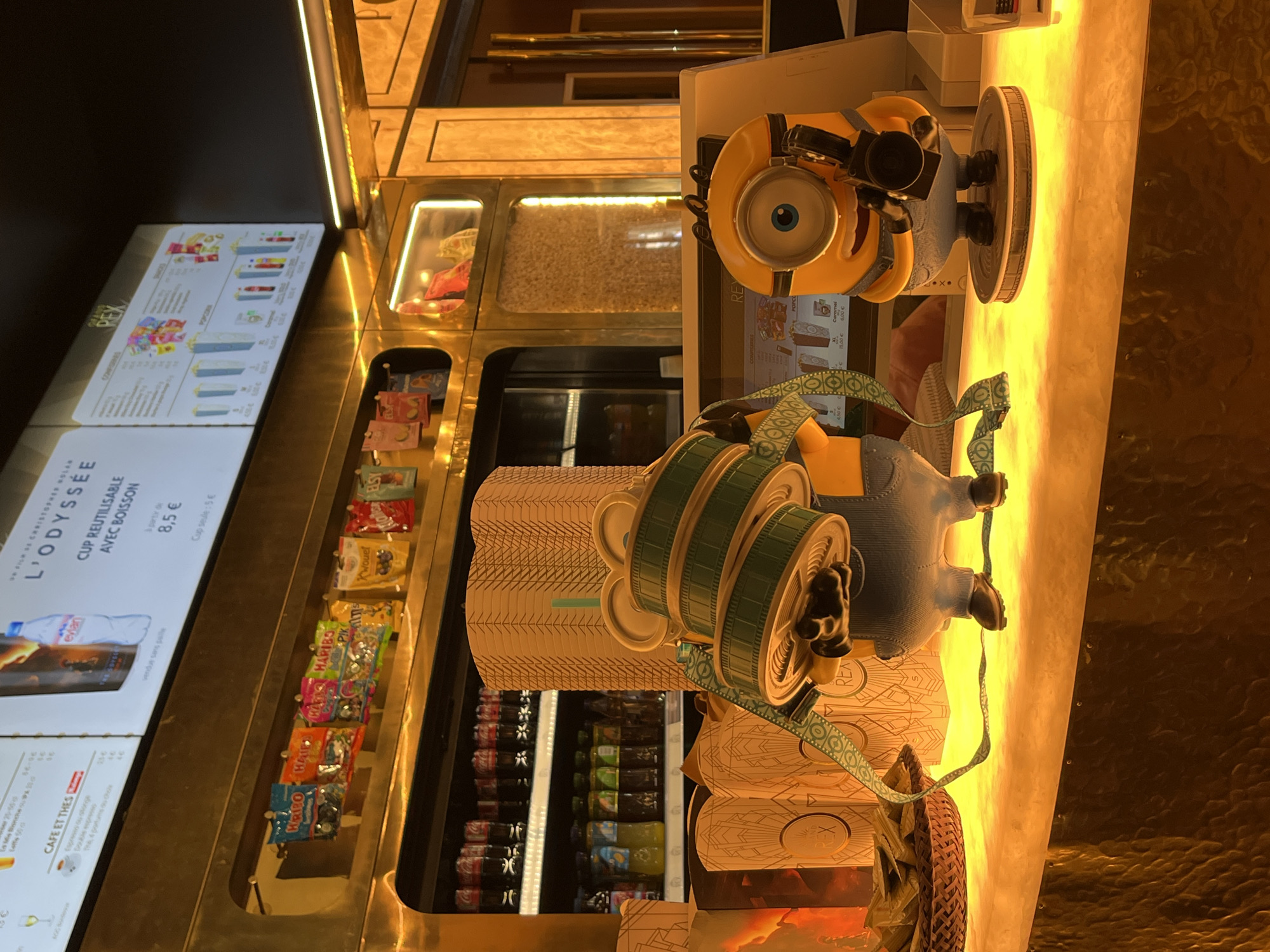 The image depicts a concession stand in a cinema lobby, likely in Paris, France, given the prominent "GRAND REX" branding visible on some of the packaging. The stand features a bright, golden, metallic decor with illuminated shelves displaying various items.
In the foreground, two Minion figurines are prominently displayed on a brightly lit counter. The Minion on the left is holding a large, green, reel-like object with a patterned strap. The Minion on the right is holding a vintage-style movie camera.
Behind the figurines, a large stack of golden-colored cardboard popcorn containers is visible. Further back, a refrigerated display case contains rows of bottled beverages, including Coca-Cola and what appear to be fruit juices. Above the refrigerated case, a digital menu board displays movie titles and prices, including "L'ODYSSÉE" and "REX". Various bags of candy, such as Haribo, are displayed on shelves to the left of the menu board.
The overall impression is one of a vibrant and well-stocked cinema concession area. The lighting is warm and inviting, highlighting the golden accents of the decor.