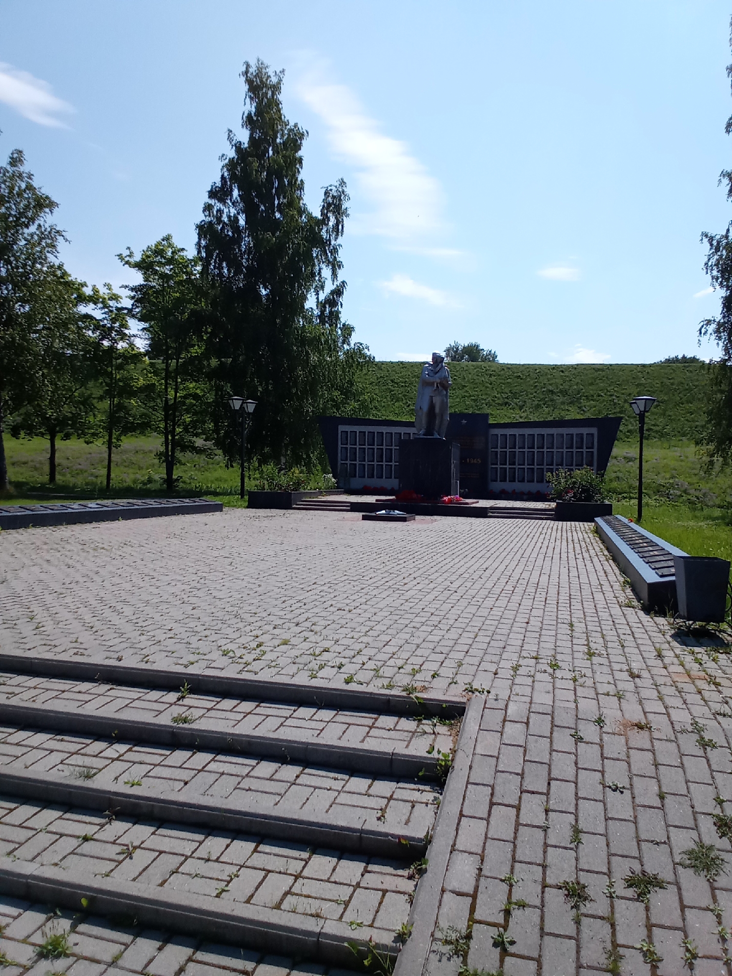 The image depicts a solemn memorial site in Belozersk, Russia, likely dedicated to war veterans. In the foreground, a paved walkway with visible grass growing between the stones leads up a set of wide steps. To the left, a long, low granite structure holds several dark rectangular plaques. The central element of the memorial is a monument featuring a statue of a soldier standing before a dark, curved wall adorned with numerous smaller plaques or portraits. A wreath of red flowers lies at the base of the monument. A streetlamp stands to the right of the central monument. In the background, a grassy embankment rises behind the memorial, topped with trees. The sky is a clear blue with a few wispy clouds. The scene is tranquil and appears to be captured on a sunny day.
