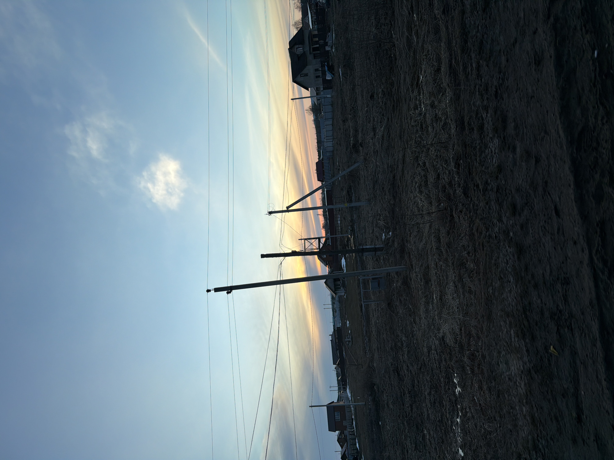 The image depicts a rural scene, likely on the outskirts of the city of Дулесово, Russia, given the presence of utility poles and sparse buildings against a backdrop of open land. The landscape is dominated by a field of dry, withered vegetation, suggesting a season like late autumn or early spring. Utility poles stand prominently in the foreground, with electrical lines stretching across the sky. A few houses are visible in the distance, their silhouettes outlined against the sunset. The sky is gradient of blues and oranges, characteristic of twilight.