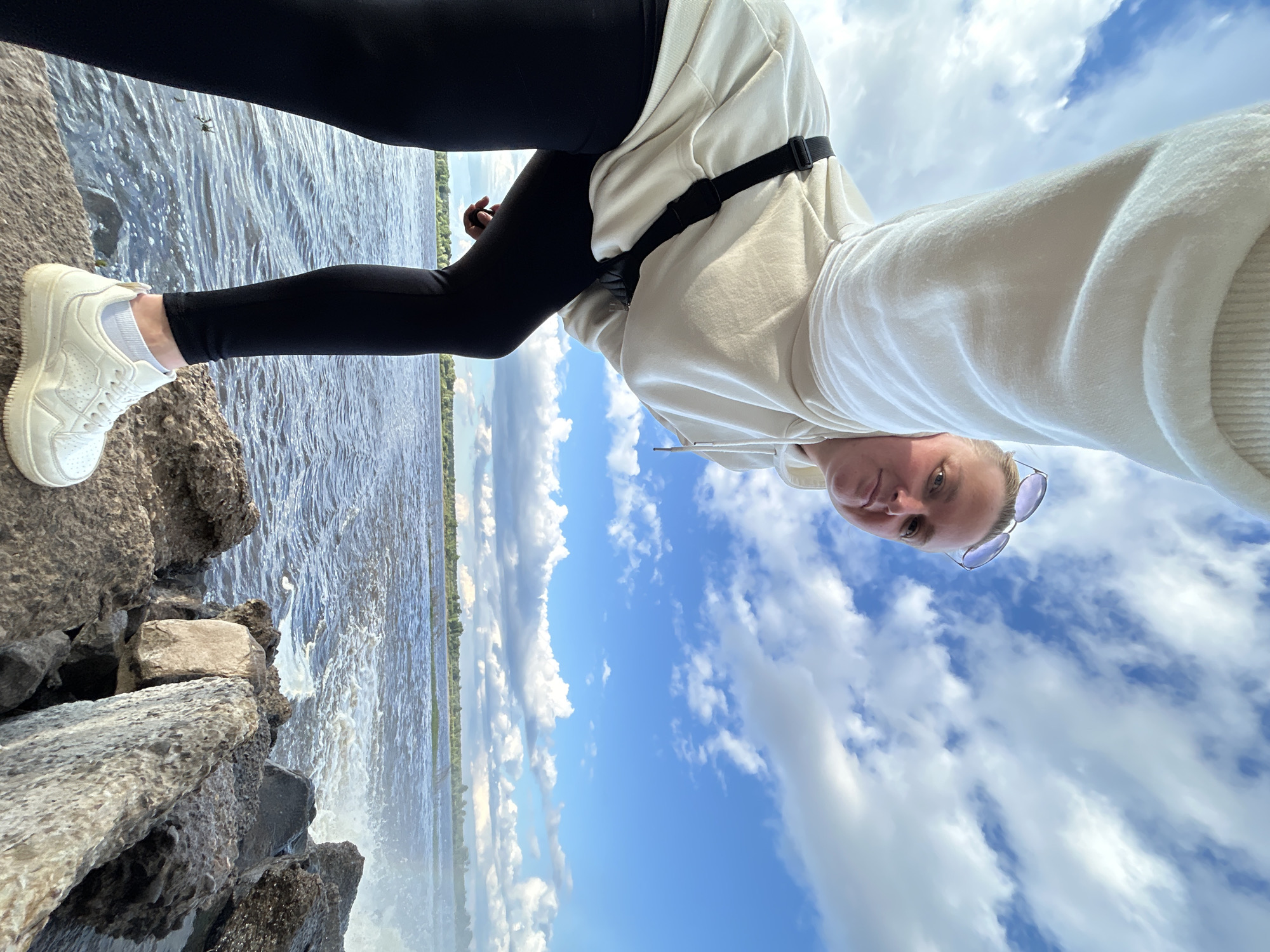 A low-angle, full-body selfie captures a woman standing on a rocky shore, with a body of water and a tree-lined horizon in the background. The woman, with blonde hair pulled back, wears sunglasses perched on her head, a cream-colored hoodie, black leggings, and white sneakers. A black fanny pack is slung across her chest. The choppy water suggests a windy day, and the sky is filled with white, puffy clouds against a bright blue expanse. The rocks in the foreground are large and varied in texture and color. The location is identified as the bank of the Kama River in the city of Chernushka, Russia.