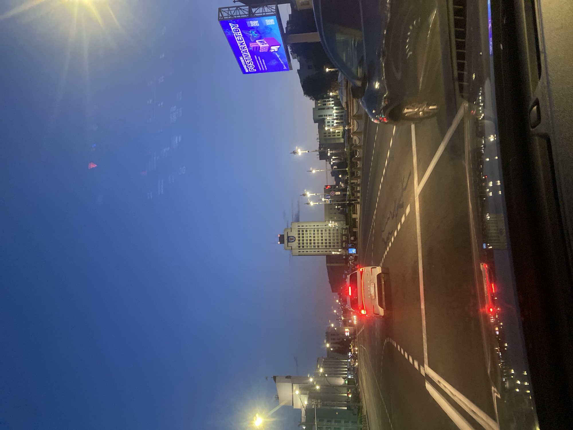 The image shows a street scene in Minsk, Belarus, at dusk. The sky is a deep blue, with the sun just below the horizon, casting a warm glow. A large billboard advertising "POSTMARKET.BY" is prominently displayed on the right side of the road. Several cars are on the road, with their headlights and taillights illuminated. The foreground shows the dashboard and windshield of a car, reflecting the city lights. In the background, modern buildings line the street, including a tall white building with a distinctive architectural style. The overall impression is of a bustling city transitioning from day to night.