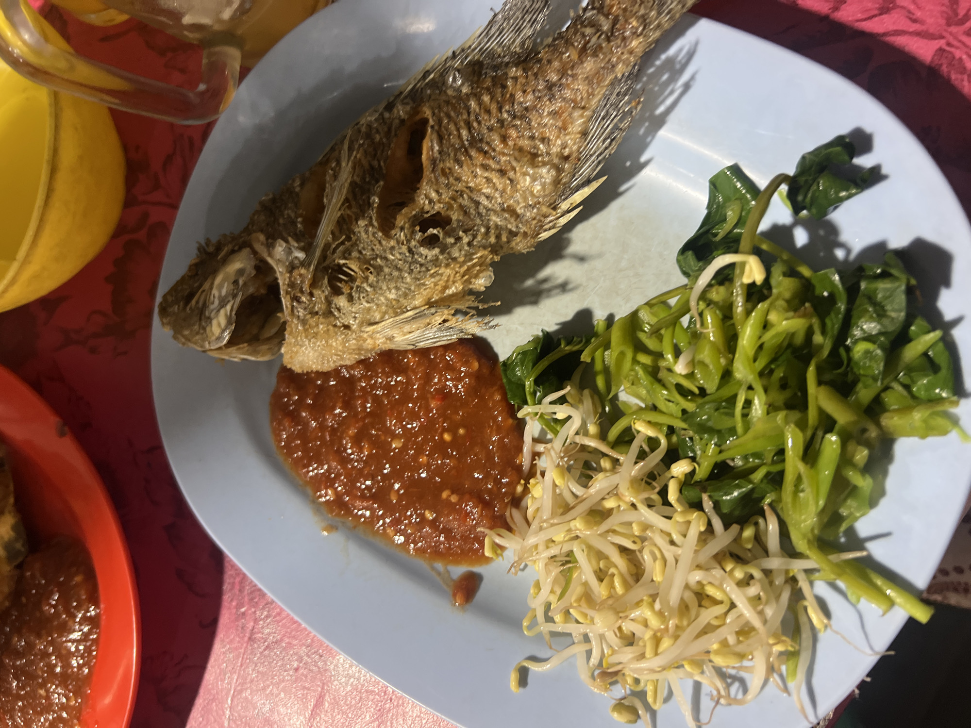 In Gianyar, Indonesia, a light blue plate holds a meal. The plate contains a whole fried fish, a scoop of red chili sauce, a mound of bean sprouts, and a pile of cooked green vegetables. A red tablecloth is visible beneath the plate. In the upper right of the frame, a glass pitcher filled with a light yellow liquid is partially visible. The scene appears to be at a food stall or a casual dining establishment.