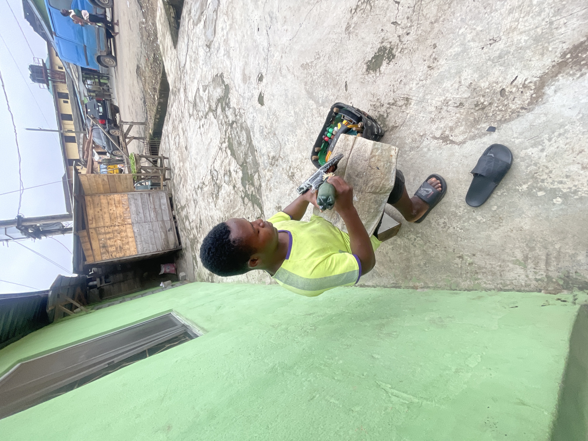 A young Black person wearing a neon yellow t-shirt with purple trim and black shorts is sitting on a concrete surface, leaning against a bright green wall. They are holding a tool, possibly a glue gun, and working on a piece of wood. Next to them is a black plastic crate filled with various items, including colorful balls, rolls of tape, and other tools. A pair of black flip-flops is on the ground nearby. The environment appears to be an outdoor urban setting in Akowonjo, Nigeria, with a wooden stall structure in the background, a paved area with some water stains, and a street with vehicles including a blue van and a black SUV. The sky is overcast.