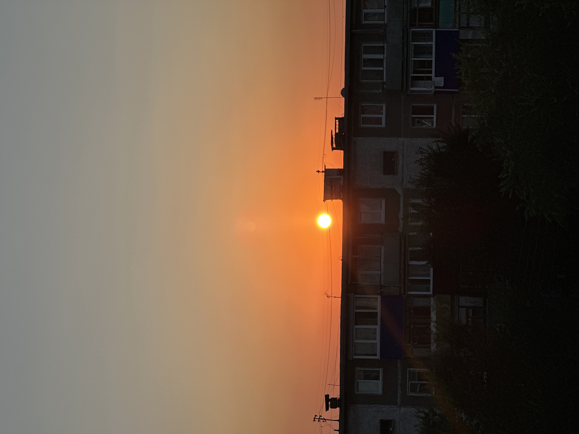 A vertical, medium-shot photograph captures a sunset over a Soviet-era apartment building in Energetik, Russia. The sun, a bright orange orb, is positioned in the upper-middle section of the frame, just above the roofline of the building. The sky transitions from a pale gray at the top to a vibrant orange near the horizon.
The apartment building, which occupies the lower half of the image, appears to be a multi-story structure with numerous windows and balconies. Many of the windows are dark, but some reflect the sunset's warm glow. On the roof, there are various structures including what appear to be ventilation shafts, chimneys, and antennas. Below the building, dense, dark green foliage obscures the lower floors and grounds. The overall impression is one of a typical urban evening scene.