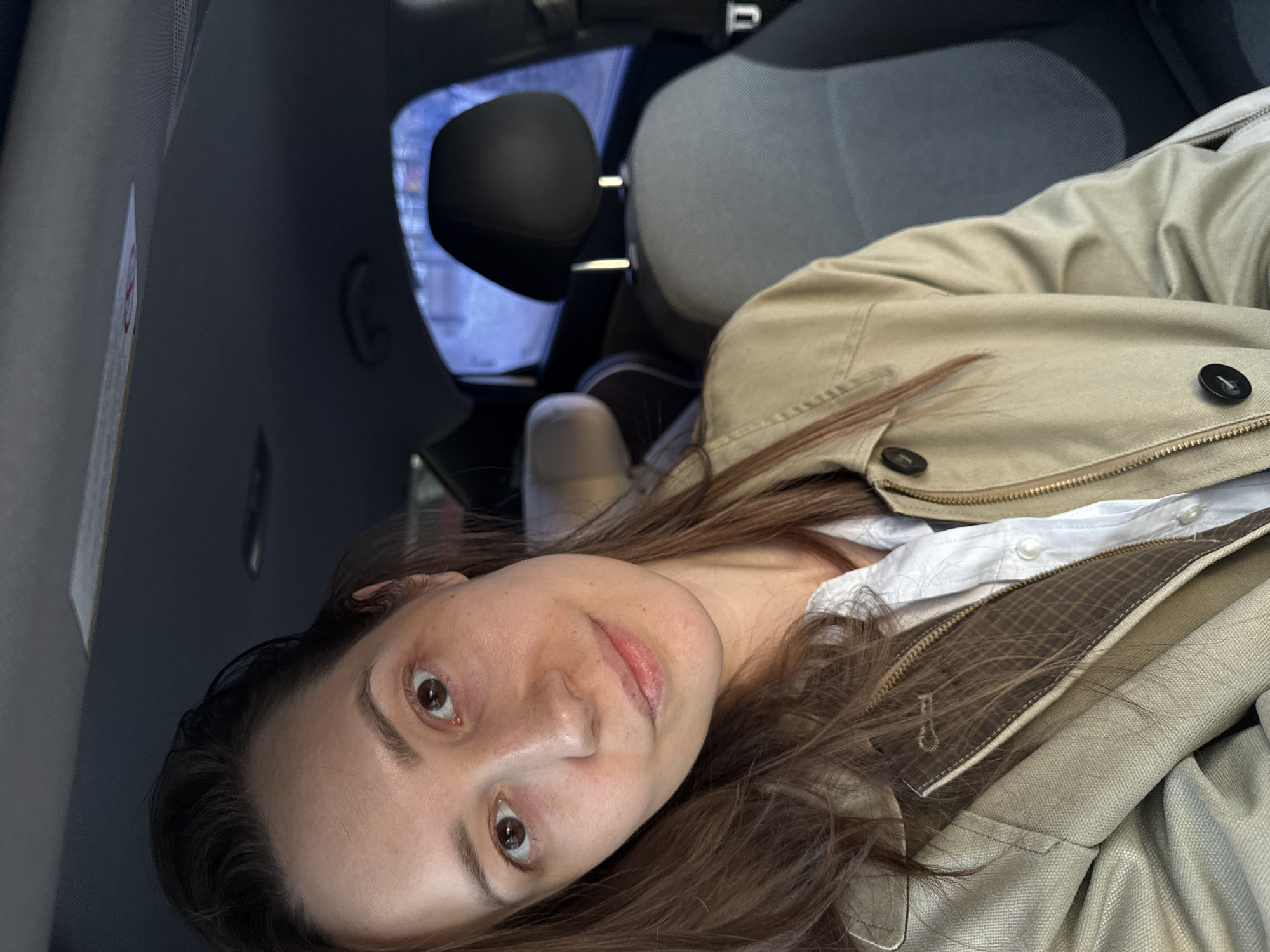 A female individual is depicted in a close-up, chest-up portrait taken inside a vehicle in Chaikovsky, Russia. She has fair skin, brown eyes, and long brown hair, and looks directly at the camera with a neutral expression. She wears a light-colored collared shirt beneath a light tan or olive-colored jacket featuring a zipper closure and dark buttons, with a visible brown plaid lining near the zipper. The vehicle's interior consists of a dark gray headliner, an overhead light fixture, and a partially visible sun visor with a sticker. To the subject's right, a black headrest and gray fabric upholstery of a rear car seat are visible. To her left, part of a front passenger seat with a lighter gray headrest and what appears to be a child seat attachment are visible. The exterior environment, viewed through the vehicle's rear window, is indistinct but suggests an outdoor, possibly urban setting under muted light, with vague colorful elements and a light-colored ground surface, potentially snow. The scene captures the individual stationary within the vehicle.