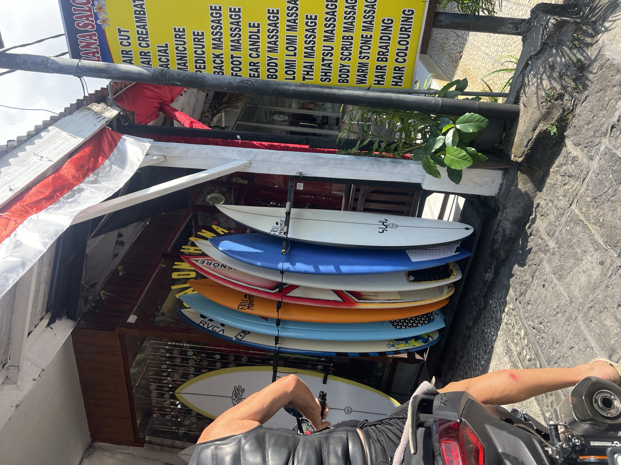 In Kuta, Indonesia, a person is seated on a motorcycle parked in front of a surf shop. The motorcycle's rear section is visible on the left, with the person's legs extending downwards. The person is wearing black shorts and a black vest. Their right leg shows a reddish abrasion. They are wearing sandals.
The surf shop displays numerous surfboards lined up vertically. The surfboards are of various colors and designs, including yellow, blue, orange, and white. Some surfboards have text and logos visible, such as "RVCA" and "FREE MOVEMENT". A white surfboard with yellow trim is positioned on the far left.
A yellow sign with black text is displayed to the right of the surfboards, listing various services such as "HAIR CUT," "MANICURE," "PEDICURE," "BACK MASSAGE," "FOOT MASSAGE," "BODY MASSAGE," and "HAIR BRAIDING." A red and white Indonesian flag is draped over the awning of the shop.
The ground is paved with stone tiles. A small potted plant with green leaves is situated near the surfboards and the yellow sign. The overall environment suggests a commercial area, likely a street or walkway, in a warm climate.