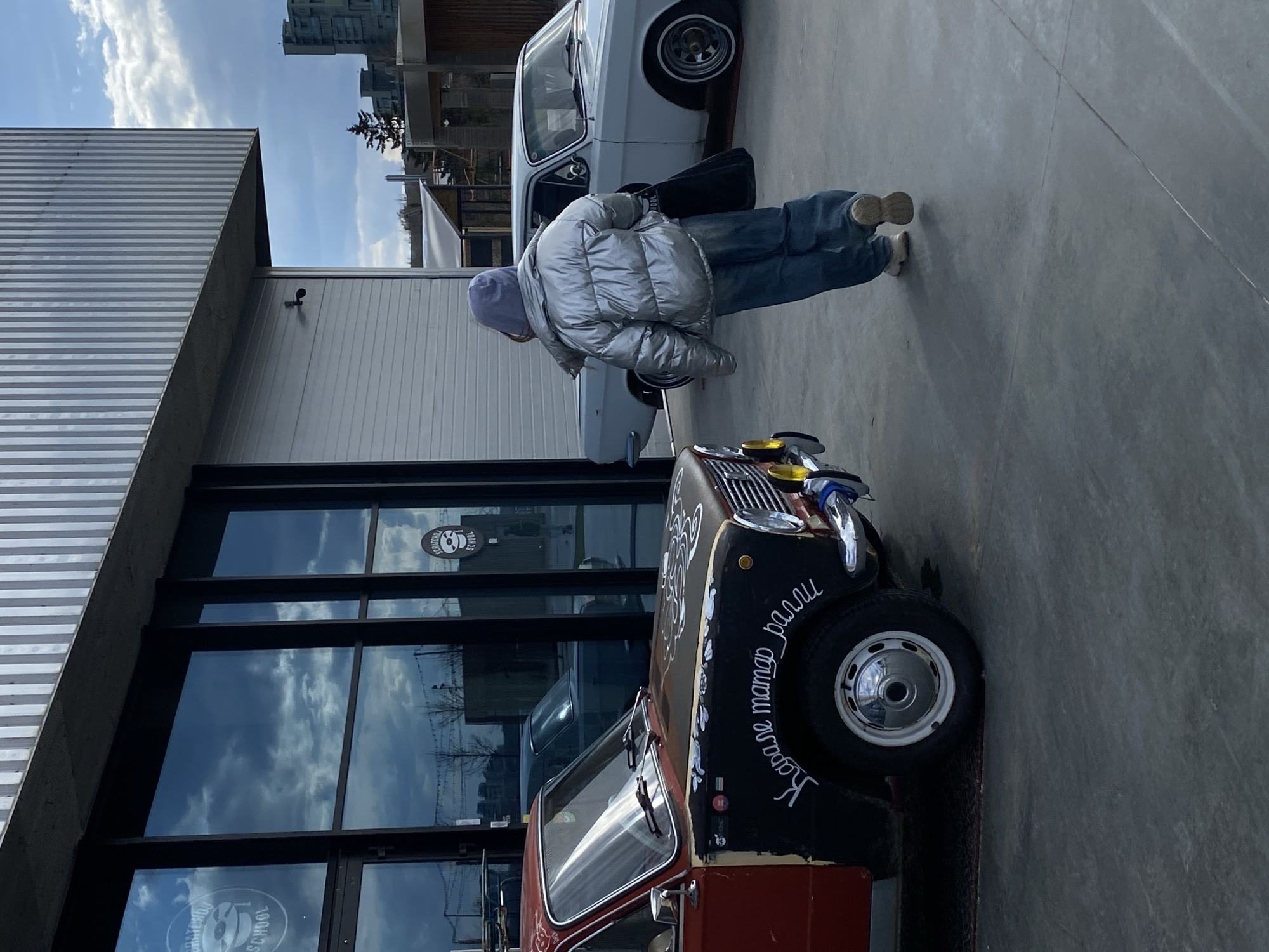 The image captures an outdoor urban scene in Kazan, Russia, on a bright, partially cloudy day.
In the foreground, a person stands with their back largely to the viewer, dressed in a shiny silver puffer jacket, blue jeans, and a light blue beanie. They are leaning slightly forward, seemingly observing a distinctive customized car parked in front of them.
This unique car, positioned in the mid-ground, features a two-tone paint scheme: black on the top and hood, and a reddish-brown on the lower body. White, stylized Cyrillic text is hand-painted on its black side, reading "Кареглазая мама ралли" (Kareglazaya mama ralli), which translates to "Brown-eyed mama rally." The vehicle has prominent chrome accents, including bumper elements and headlight surrounds, and yellow-tinted auxiliary lights on the front, suggesting a custom or vintage rally car, possibly a classic Mini. Behind the person, a light-colored, possibly white or light grey, vintage sedan is partially visible.
The scene is set on a light grey concrete or paved surface. To the left, a building with a silver corrugated metal facade is seen under a concrete overhang. To the right, a building features large glass windows that reflect the blue sky and clouds. Several round stickers are visible on these windows; one clearly says "SCRATCH SCHOOL" and another appears to be a "SOUNDFACTORY SCHOOL" sticker, possibly indicating a music or DJ school. In the distant background, tall urban buildings and some scattered trees are visible against the sky. The overall impression is an artistic, personalized car culture scene within an urban setting.