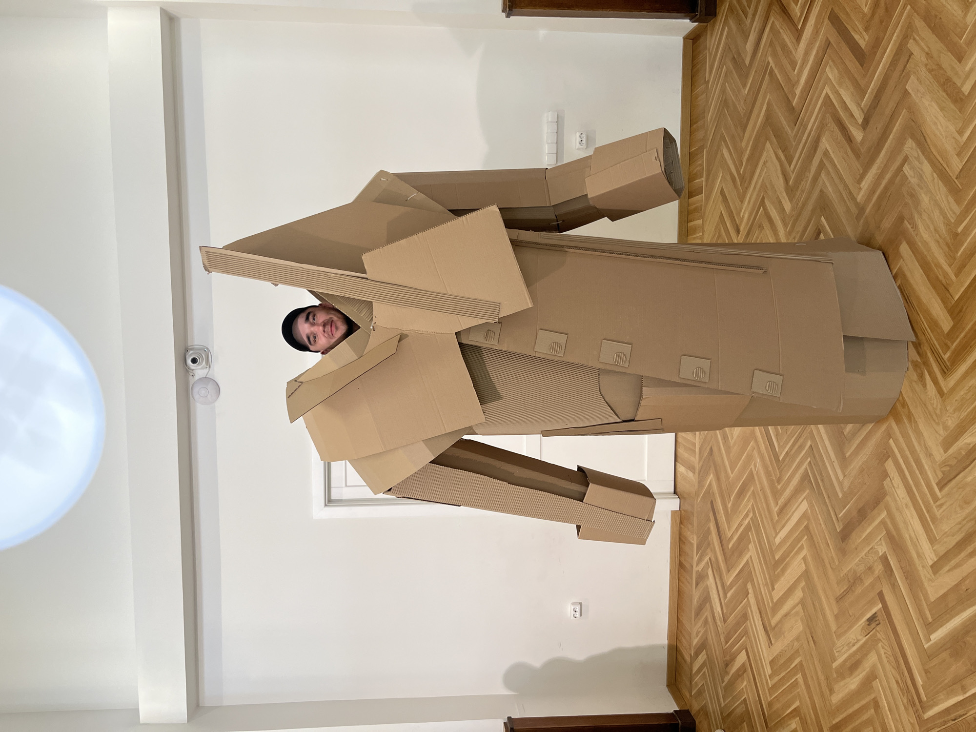 A man wearing a black beanie is encased in a cardboard robot costume, standing in a room with light-colored walls and a parquet wooden floor. He is positioned in the center of the frame, facing the camera with a slight smile. The cardboard costume is complex, with angular shapes and protruding elements resembling robot limbs. A white door and electrical outlets are visible on the wall behind him. A large, round light fixture is visible on the ceiling in the upper left corner. The location is identified as being in Yekaterinburg, Russia based on other context that is not present in the image.