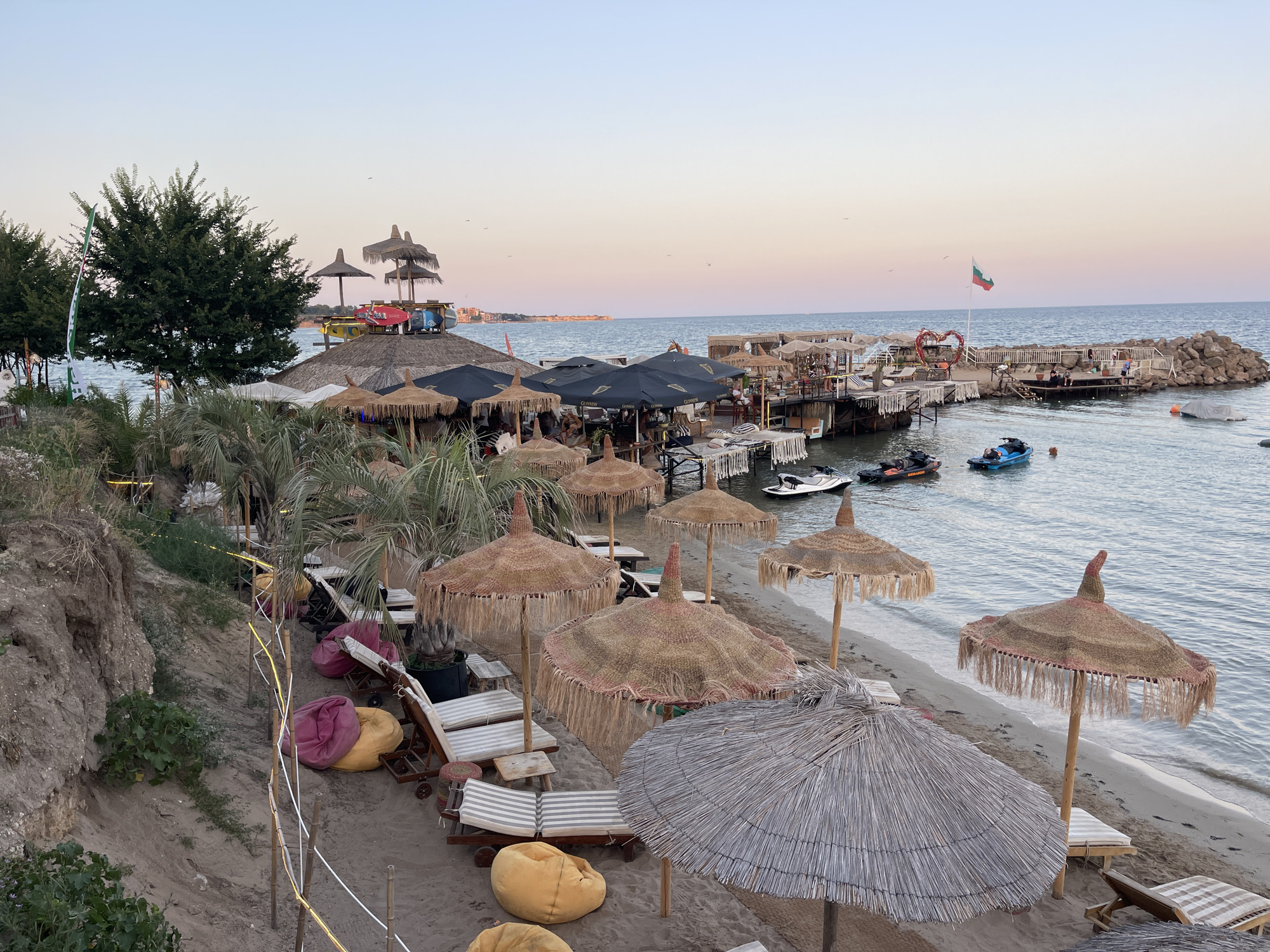 This image depicts a beachside area in Ravda, Bulgaria, with a clear sky at dusk. The scene is characterized by a sandy beach populated with numerous sun loungers and beanbag chairs, many of which are unoccupied. Several thatched umbrellas, some with fringed edges, provide shade. In the background, a pier-like structure extends over the water, featuring more seating areas and umbrellas, some of which are dark-colored with prominent branding, suggesting a restaurant or bar. Watercraft, including jet skis, are visible in the water near the pier. A Bulgarian flag flies from a flagpole on the pier. The overall atmosphere is tranquil, with the setting sun casting a soft light over the scene. The presence of a rocky embankment on the left side of the image indicates a slight elevation change.