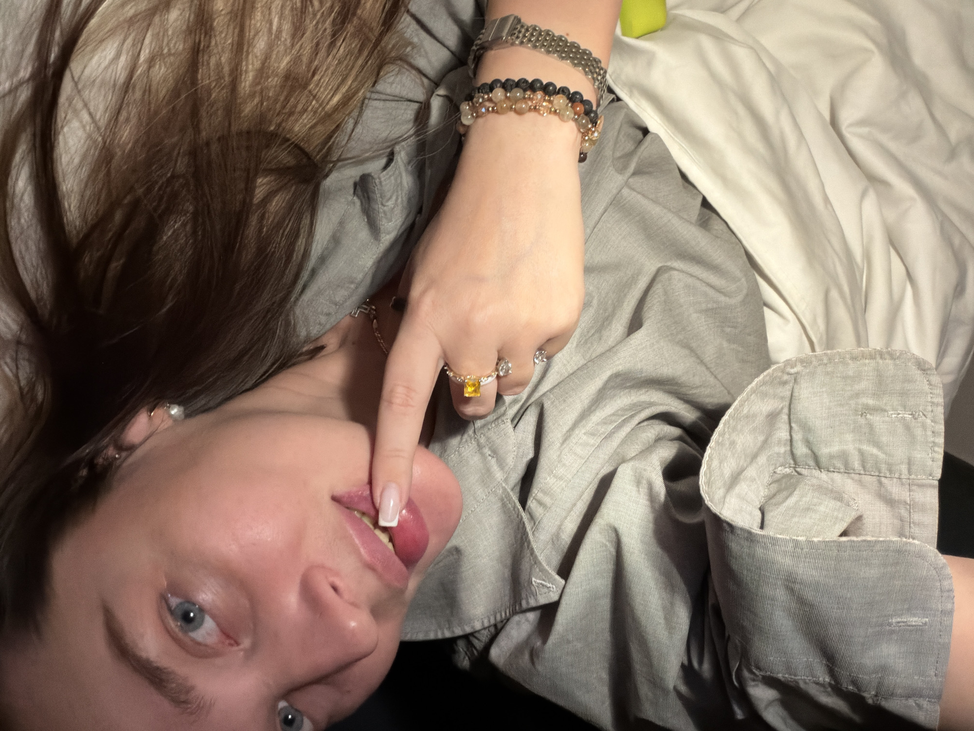 This media file presents a close-up, rotated view of a person lying down, looking directly at the camera. The individual has long brown hair, light skin, and blue eyes. They are wearing a light gray collared, long-sleeved shirt.
On their right hand, they wear several pieces of jewelry: multiple rings, one prominent with a rectangular yellow stone and another with clear sparkling stones. Their right wrist is adorned with an assortment of beaded bracelets in earthy tones (including black and amber-like beads) alongside a silver-toned metal watch. A French manicure is visible on the fingernail of the right index finger, which is playfully placed on the lips, slightly parting them. A small pearl earring is visible in their left ear.
The background primarily features crumpled white fabric, likely bedding, on the right side, and a darker, indistinct surface towards the left and bottom. A hint of a bright green object can be seen in the top right corner of the frame. The lighting is soft and even, suggesting an indoor setting. The direct gaze combined with the finger gesture creates a playful, intimate, or suggestive mood. There is no visible text in the image. The location context for this image is Moscow, Russia, though no specific geographical cues are visible within the frame.