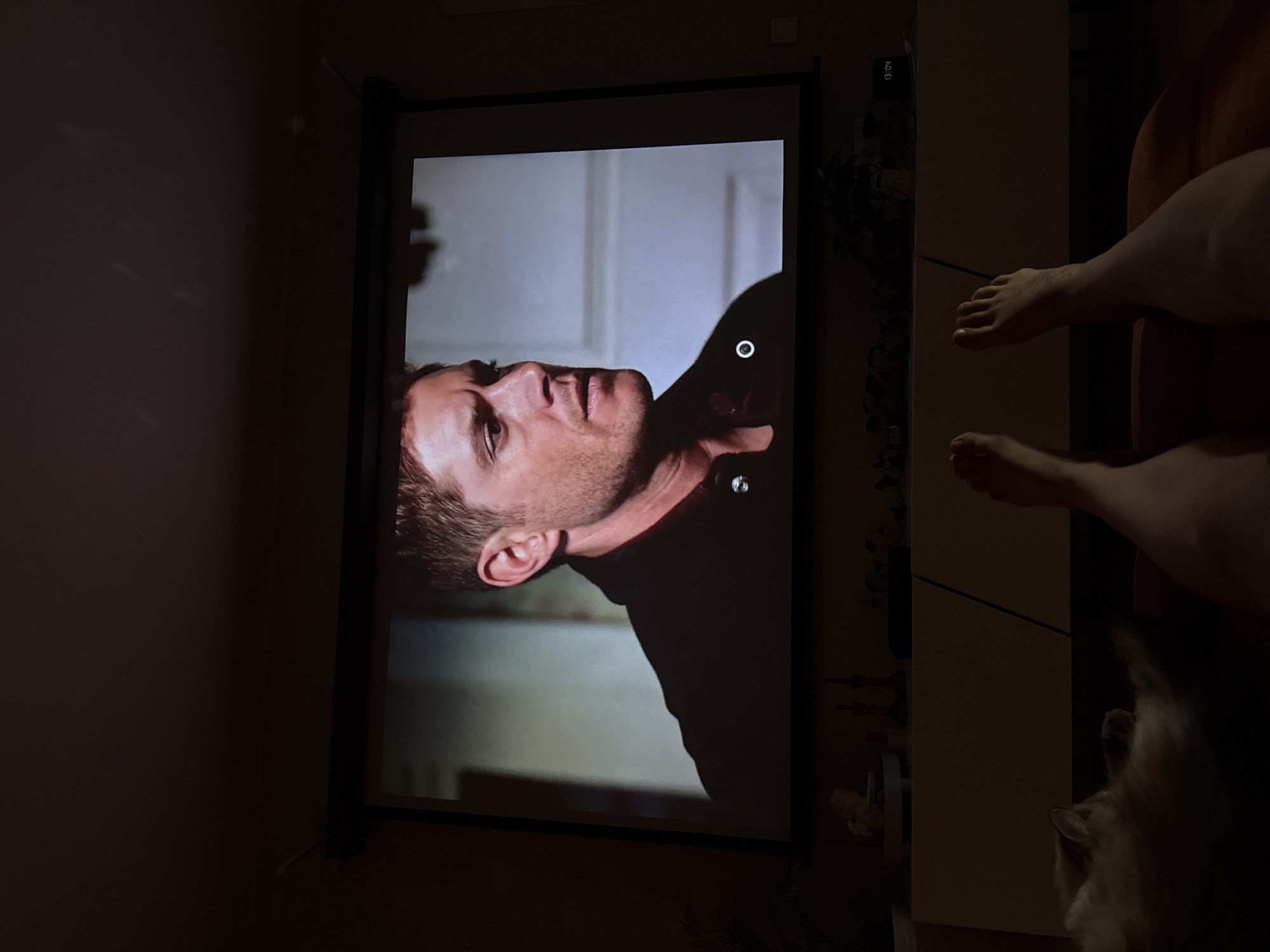 The image shows a person watching a movie projected onto a screen in a dimly lit room. The projection displays a close-up of a man's face and upper body, wearing a dark jacket. In the foreground, two feet are visible, suggesting the viewer is lying down. A white and gray cat is also present, partially visible on the left side of the image, seemingly asleep or relaxed. The background features a cabinet with several decorative items and plants, as well as a digital clock displaying "13:04". The scene appears to be set in a residential apartment in Rostov-on-Don, Russia.
