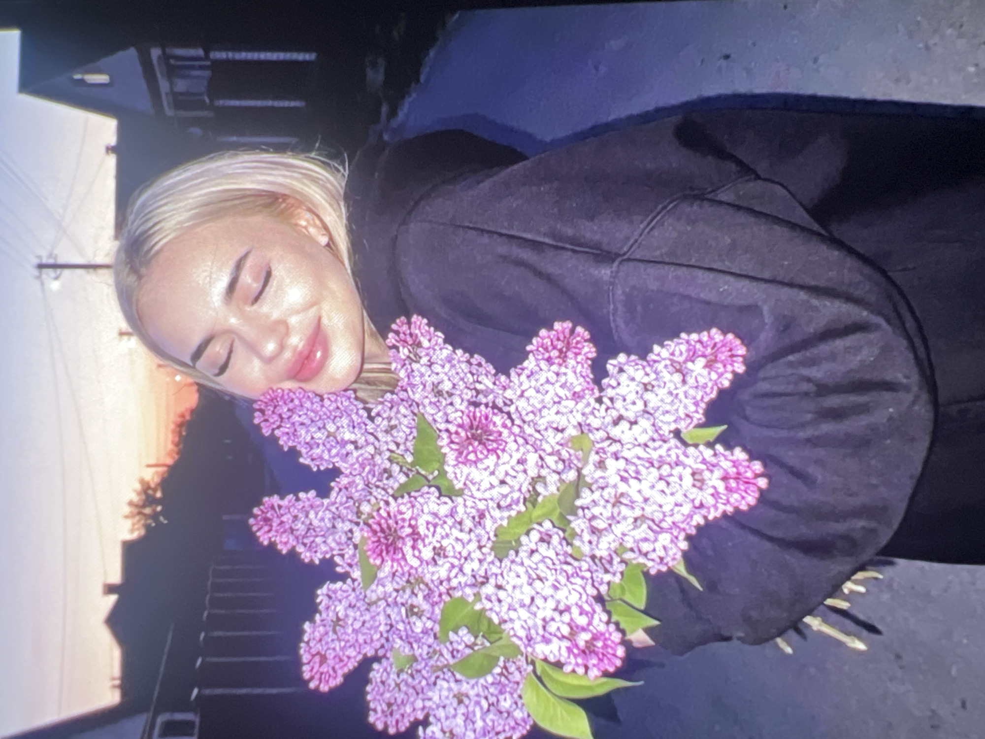 A young woman with blonde hair, appearing to be Caucasian, is captured in a close-up outdoor portrait. She is wearing a dark, possibly black or navy blue, hooded sweatshirt. Her eyes are gently closed, and a serene smile adorns her face, suggesting a moment of peace, happiness, or contentment. Her lips are full and pink.
She is holding a large, fragrant bouquet of purple lilac flowers, which are held close to her face, implying she might be smelling them or simply enjoying their presence. The flowers consist of numerous small, light to medium purple blossoms, with some green leaves interspersed throughout the bouquet.
The setting appears to be outdoors during either dusk or dawn, indicated by the gradient sky. The horizon displays soft orange and pink tones that fade upwards into a darker blue or purple. In the background, the silhouette of a building, possibly a house or other structure, is visible on the left, along with faint utility lines. The overall impression is one of quiet joy and appreciation for nature. No visible text is present in the image.