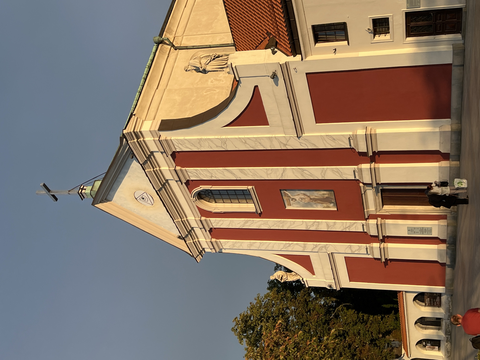 The image shows the exterior of the Church of the Holy Spirit in Lublin, Poland, bathed in the warm light of what appears to be late afternoon or early evening. The church facade is predominantly painted in a deep red with white architectural details, including fluted pilasters and cornices. A triangular pediment crowns the central section, featuring an eye within a triangle symbol and a cross at its apex. A statue of a bearded man in a robe stands on a ledge on the right side of the facade. To the left of the church, a large tree with autumnal foliage is visible.
In the foreground, a paved area suggests a public square or plaza. On the far left, a person wearing a red shirt is seated with their back to the camera. Towards the center-right of the image, two individuals, one in a dark coat and the other in lighter clothing, are walking away from the viewer.
Adjacent to the main church building, a white structure with a red tiled roof is attached, featuring a dark wooden door and two windows on its upper level. A series of arched openings, possibly part of a wall or an adjacent building, are visible on the far left, with faint signage or displays within them. The sky is a clear, pale blue, with a subtle gradient towards a slightly darker hue in the upper left corner.