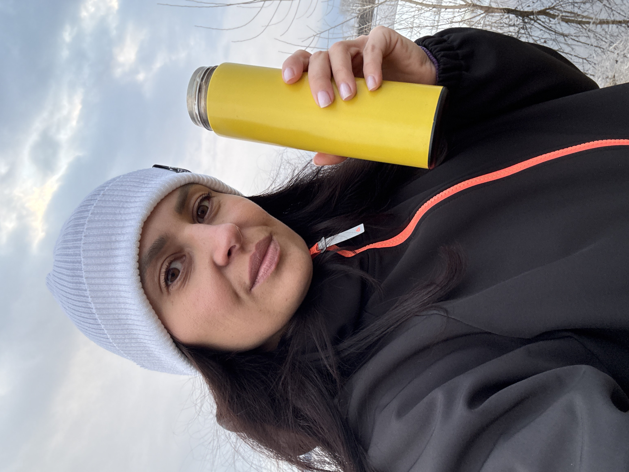 A woman is holding a yellow thermos in her right hand. She is wearing a white ribbed beanie, a black jacket with a bright orange zipper. The background shows bare trees against a cloudy sky. Given the user's request, the location is potentially in the city of Chaikovsky, Russia.