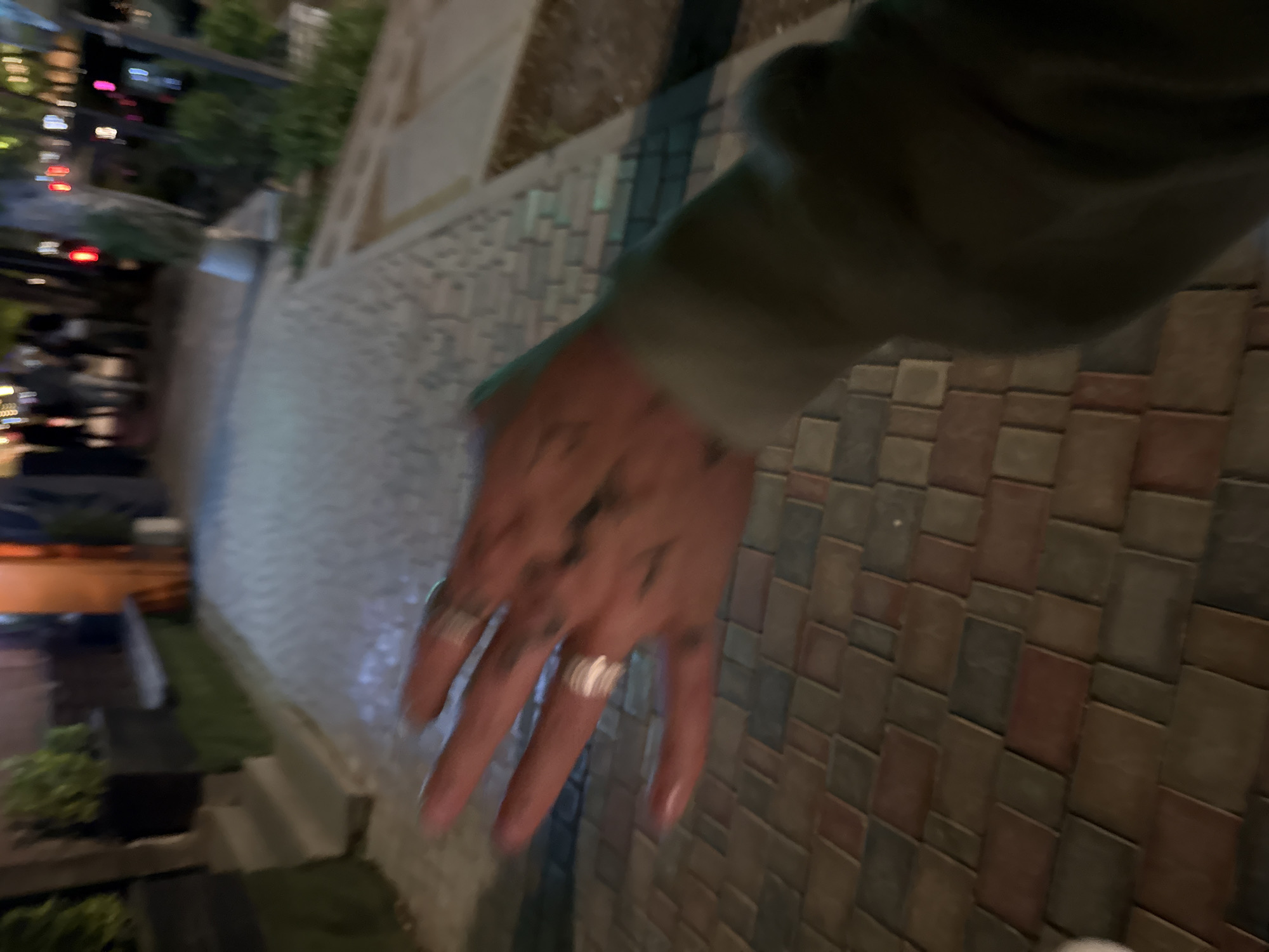 The image presents a rotated, slightly blurry, first-person perspective of an urban setting at night in Yerevan, Armenia. The main subject is a person's outstretched right hand, prominent in the foreground. This hand is adorned with two silver rings on different fingers and features distinct dark, possibly tattooed, markings across its back. The individual appears to be wearing a dark, long-sleeved garment, likely a jacket or sweatshirt, visible on the arm.
The hand extends over a paved ground surface, composed of rectangular bricks or tiles in mixed shades of brown, grey, and reddish-brown, arranged in a horizontal and vertical staggered pattern, suggesting a sidewalk or pedestrian area. The background on the left reveals blurred artificial lights, including streaks of red and white from vehicles, indicative of a busy city street. To the right, a light-colored, possibly textured wall or building facade is visible, with some dark green foliage at its base. The scene conveys a casual evening moment, possibly during a walk, in a lively urban environment. No discernible text is present in the image.