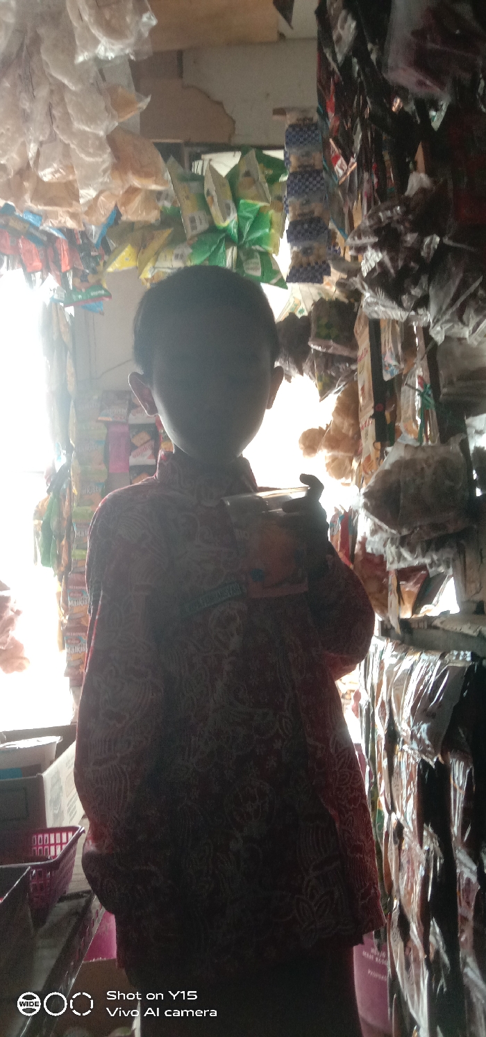 The image depicts the interior of a retail establishment, likely a small shop or stall, located in Sebaung, Indonesia. A child is positioned centrally in the foreground, silhouetted by strong backlighting emanating from an opening behind them. The child is wearing a long-sleeved red and white patterned shirt, possibly a uniform, with an illegible green badge affixed to the left chest area. They are holding a clear plastic container in their right hand. The background is densely populated with various packaged goods, consistent with snack foods or small grocery items, suspended from the ceiling and displayed on shelves. These include translucent plastic bags containing light-colored items on the left, and numerous colorful, individually wrapped packages, some green and others with a blue checkered pattern, on the right. A red plastic basket and a cardboard box are partially visible on a surface in the lower-left. The illumination causes high contrast, obscuring facial features of the child and under-exposing portions of the shop's interior.