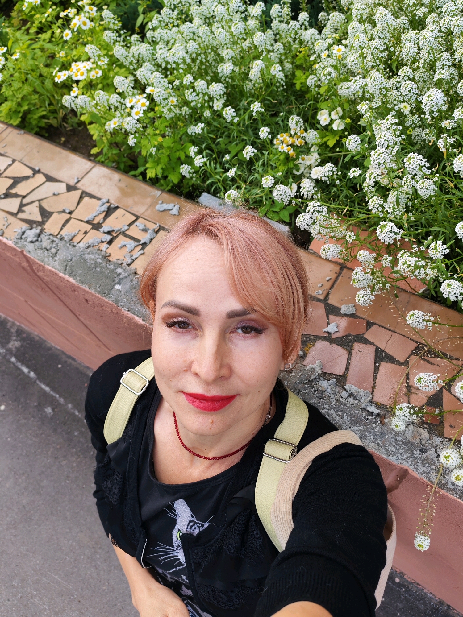 A woman with short, pink-tinted hair, wearing a black top and a red beaded necklace, is standing outdoors near a flower bed with white and yellow flowers. She is wearing bright red lipstick and has makeup on her eyes. A light-colored strap from a bag or backpack is visible across her shoulder. The background includes a decorative tiled wall and pavement. The image appears to have been taken from a high angle. The woman is likely in Chaikovsky, Russia, given the context provided.